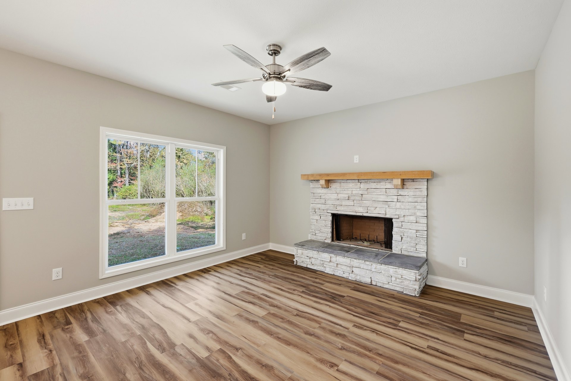 Living room with hardwood floors, white fireplace featuring a wood mantel and metal accents, ceiling fan with light fixture, large window overlooking trees.