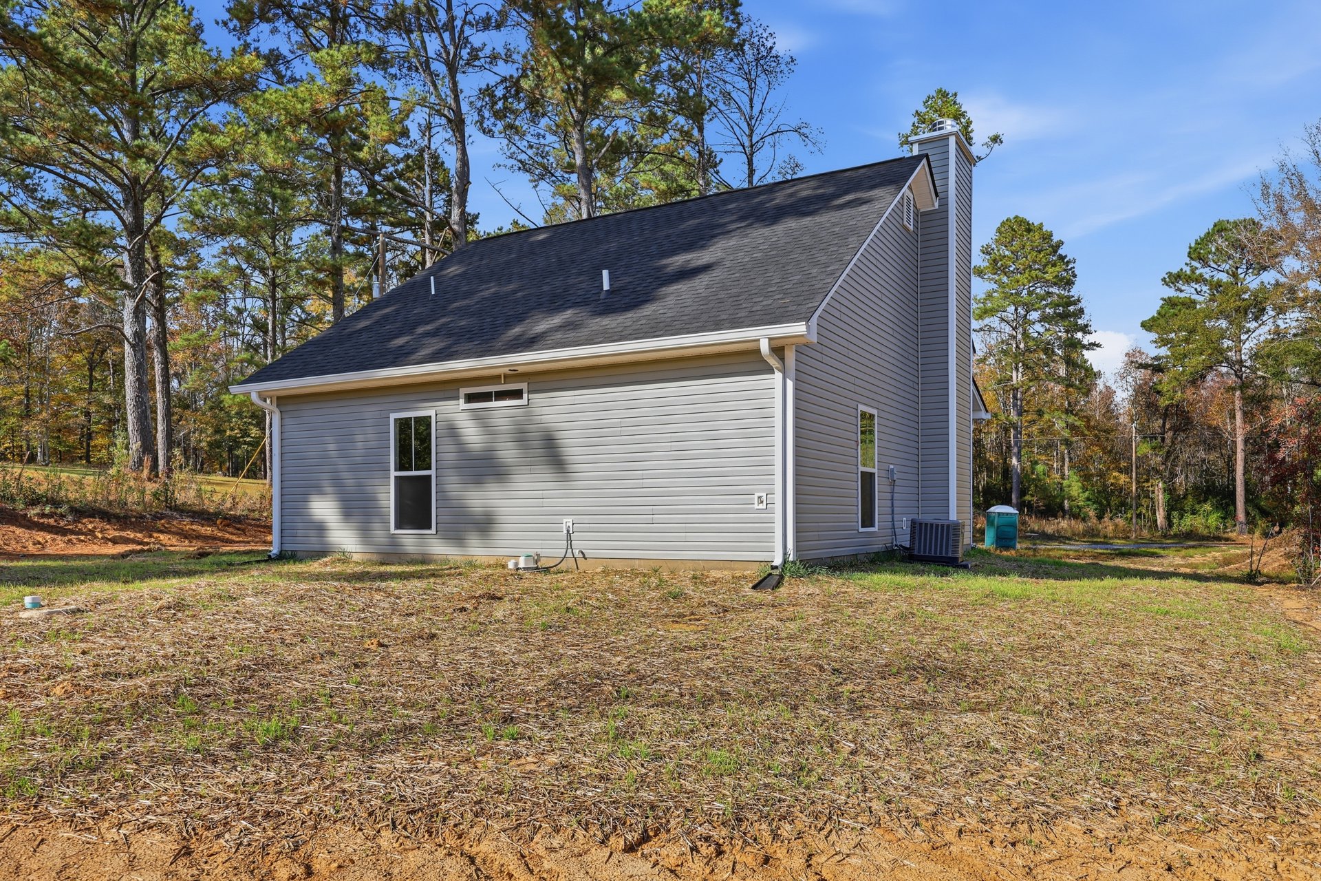 White house with a rear chimney, surrounded by grass and trees, white-framed window, black and silver air conditioner unit, hose attached to exterior wall
