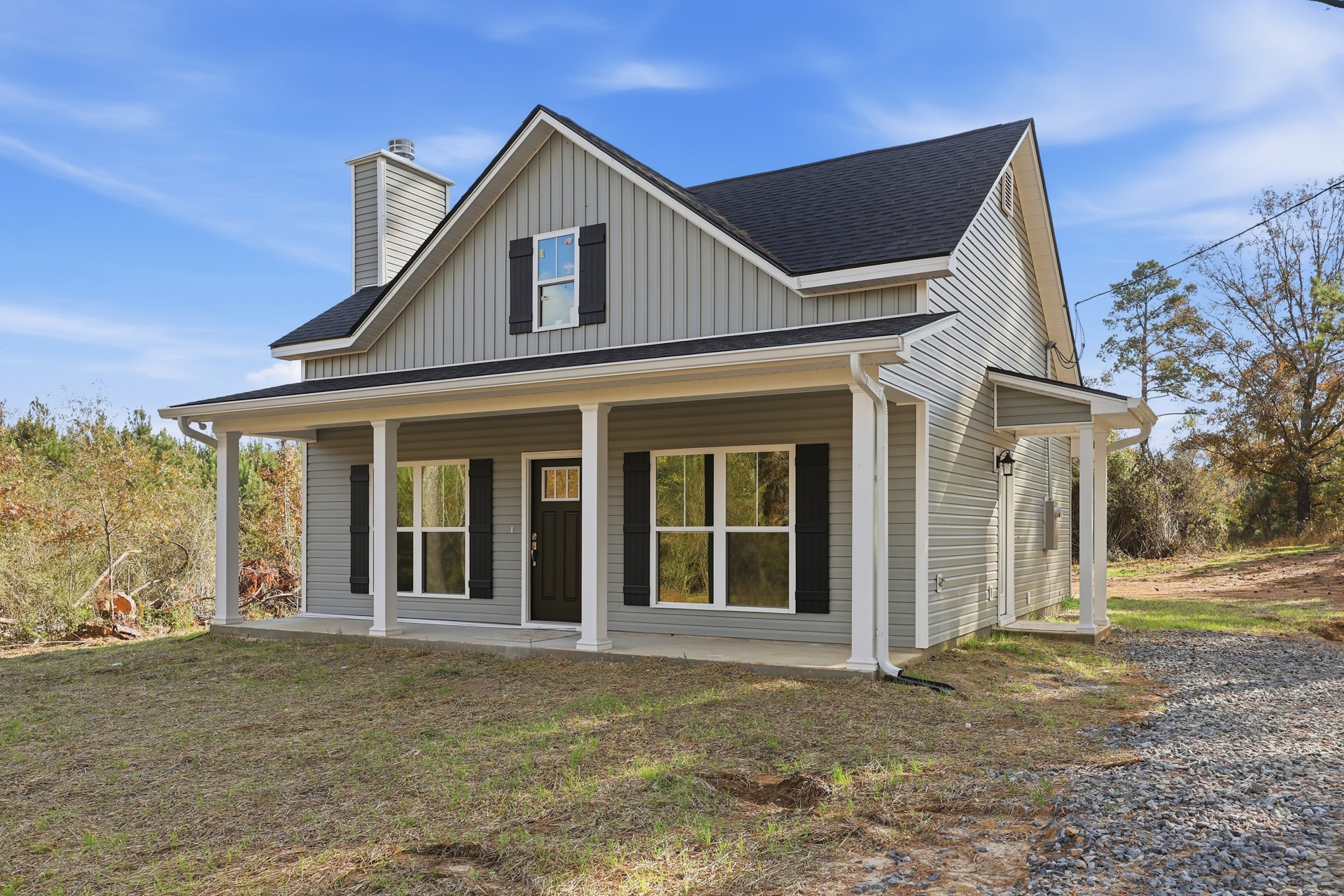 White-framed windows reflecting trees, black front door, covered porch with wood columns, gray roof, trimmed lawn with tree stump, blue sky and scattered clouds