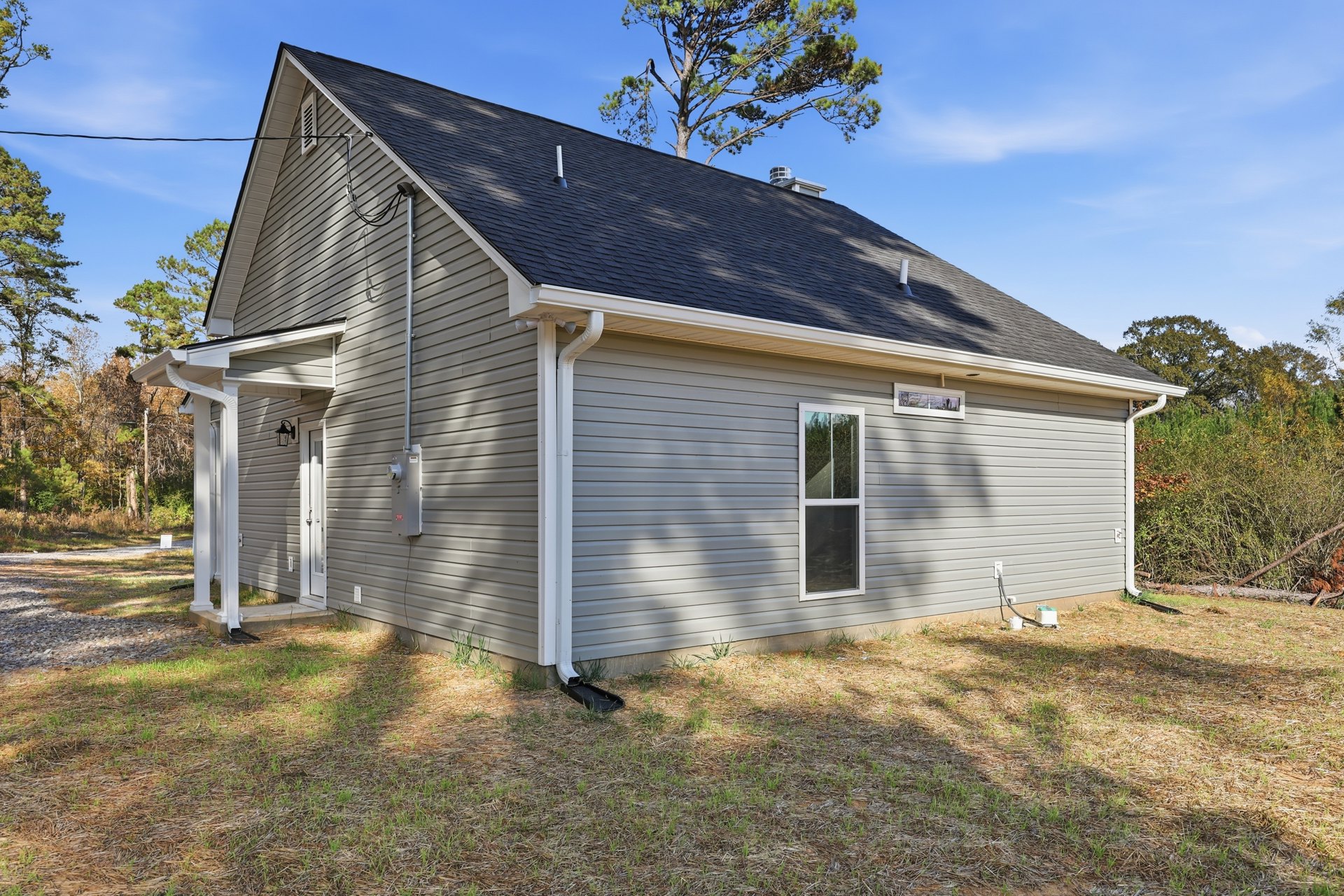 White cottage-style home with dark roof, front yard bordered by green grass and mature trees, visible window and exterior light fixture, gutter along roof edge, American Gothic