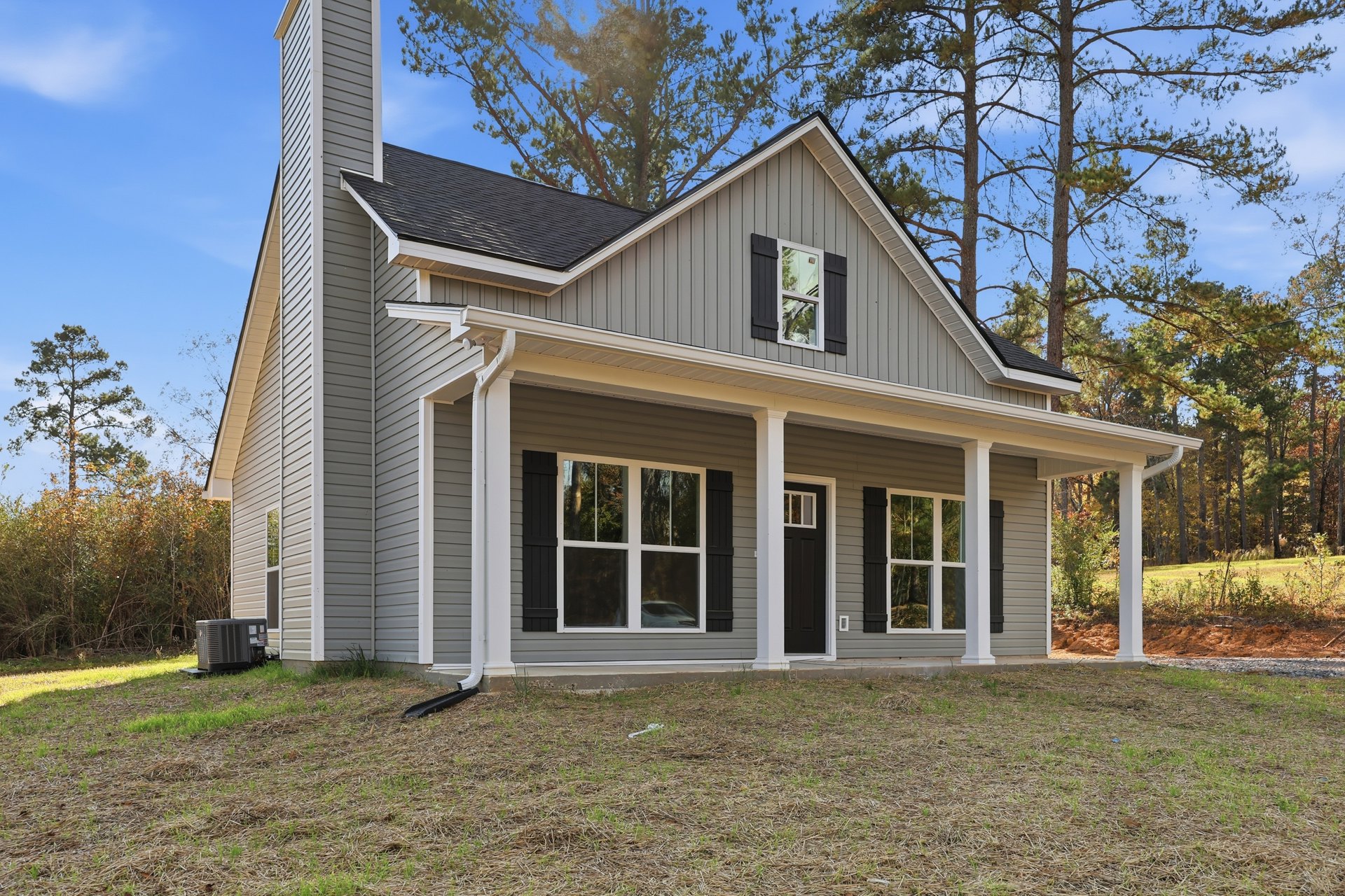Two-story house with gray siding, brick chimney, white-framed windows including one with broken glass, heat pump unit near foundation, grassy yard with white pole, mature trees in