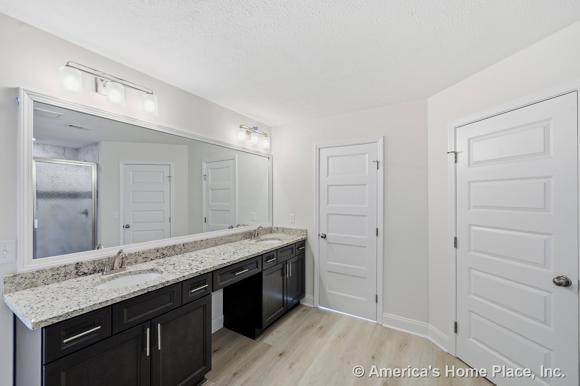 Long granite bathroom countertop with dual sinks, dark wood cabinetry, expansive wall mirror, modern vanity lighting, white paneled doors, and light wood flooring.