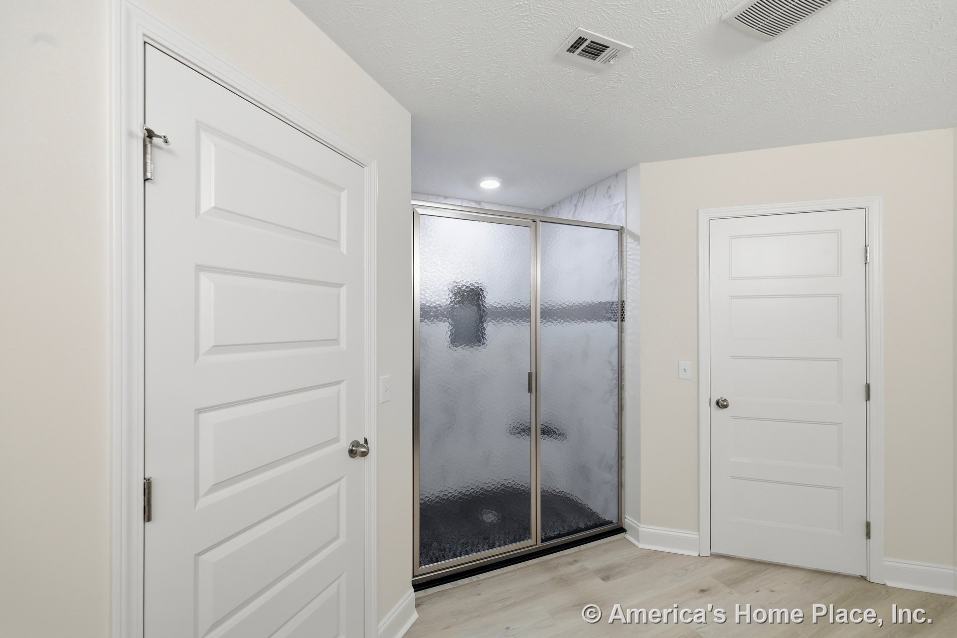 Glass-enclosed shower with textured marble-look wall tile, two white paneled doors, recessed ceiling light, light wood flooring, white baseboards, ceiling vent, and neutral walls.