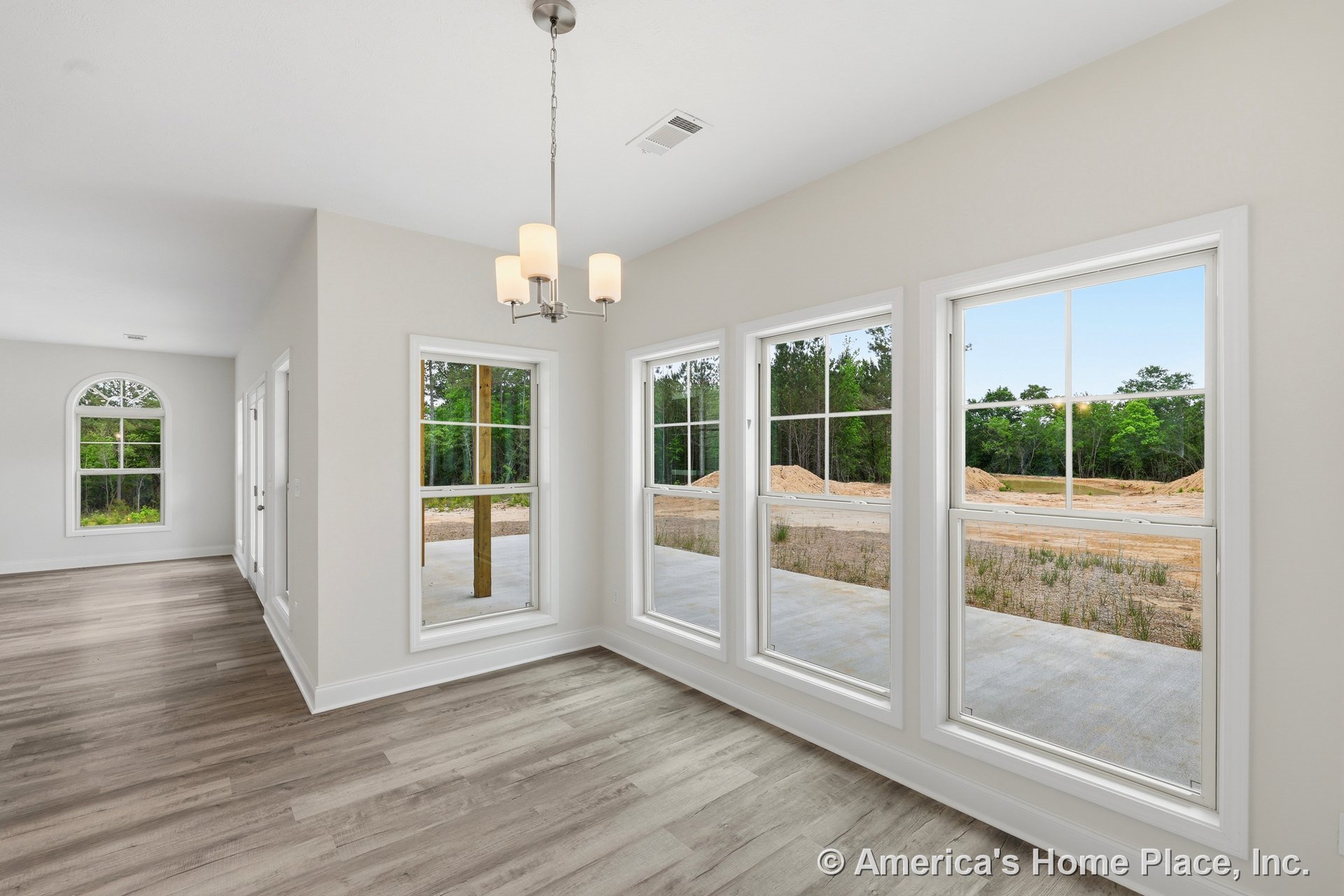 Dining area with triple picture windows, pendant chandelier, light wood plank flooring, white trim and baseboards, neutral wall color, and arched window visible in adjacent room.