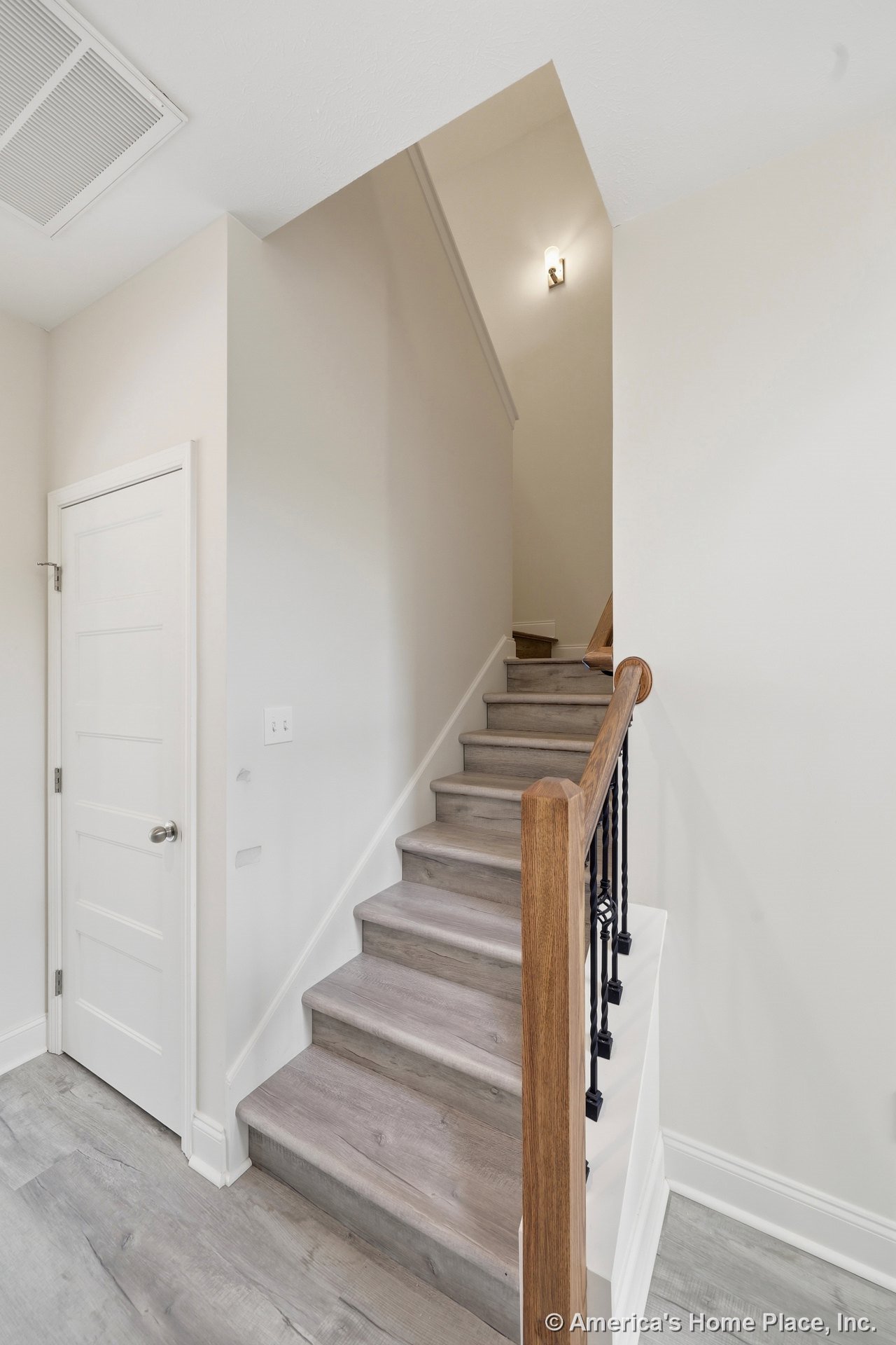 Staircase with wood-look treads, wooden handrail, black iron balusters, white trim and baseboards, white paneled interior door, wall-mounted sconce lighting, and neutral walls in a