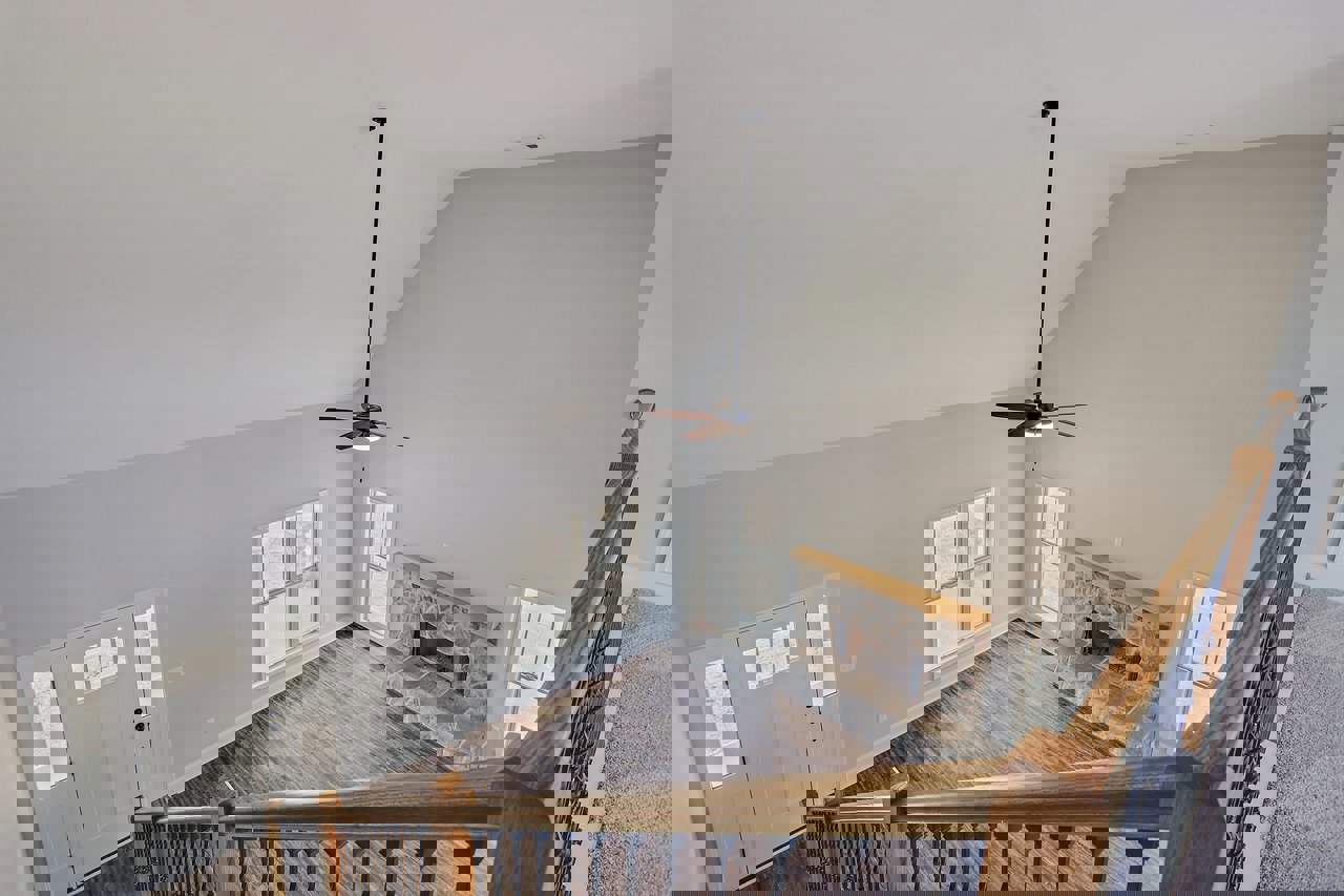 Staircase view overlooking living room with hardwood floors, white-framed window, white door with glass panes, stone fireplace, ceiling fan, and black handrail in the corner.