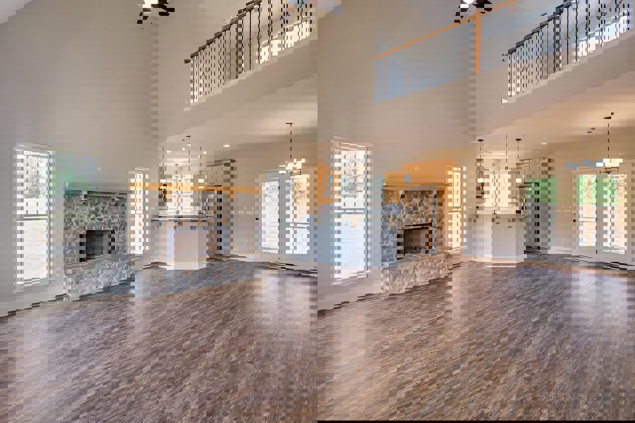 Open-concept living room with hardwood floors, stone fireplace, glass double doors, kitchen area, and large window overlooking road and trees