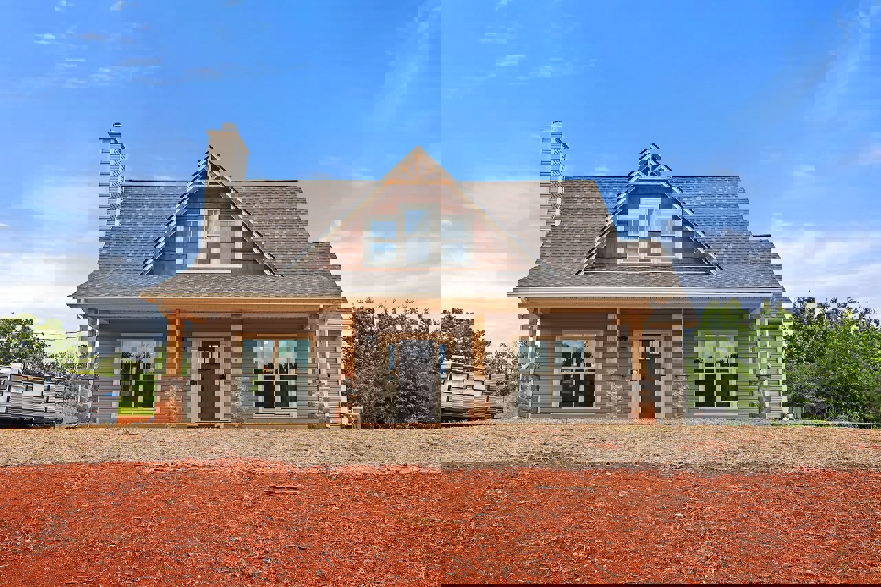 Single-story house with brown door and glass windows, surrounded by red and brown dirt yard, trailer parked nearby, scattered trees, and blue sky with clouds