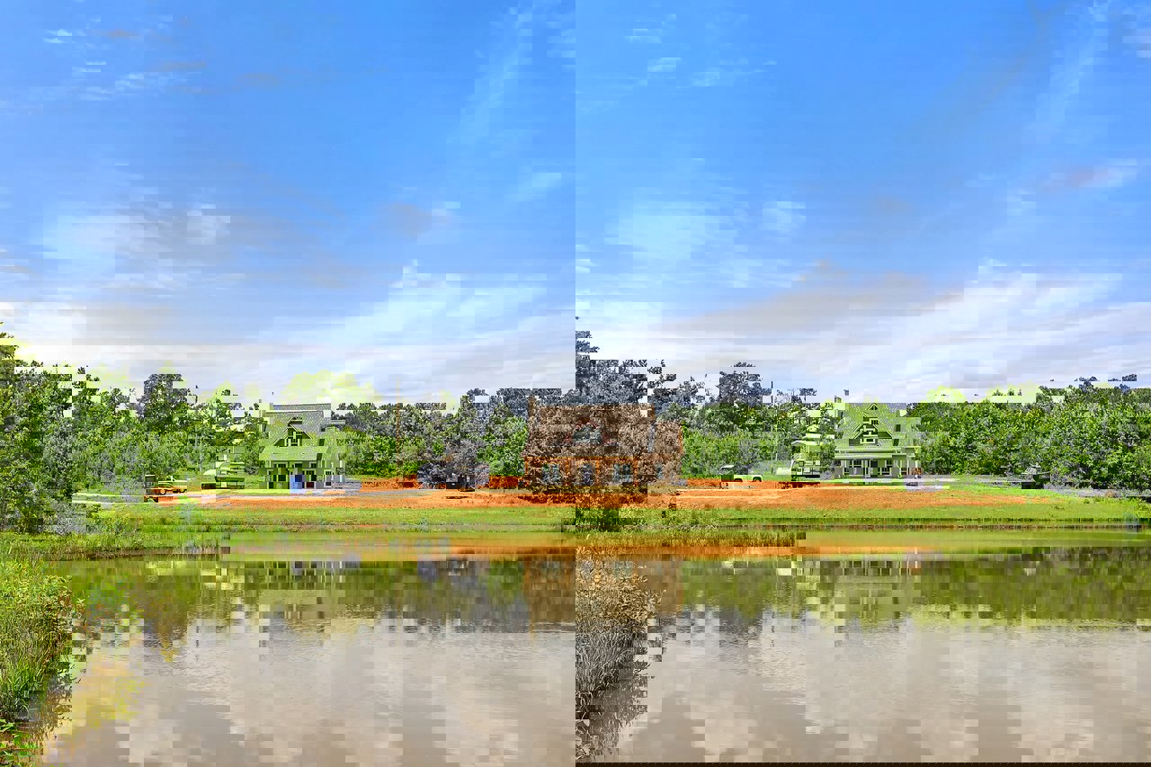 Modern lakeside home with gray roof, large windows, and wood siding, surrounded by green grass and trees, reflected in calm pond under blue sky with scattered clouds