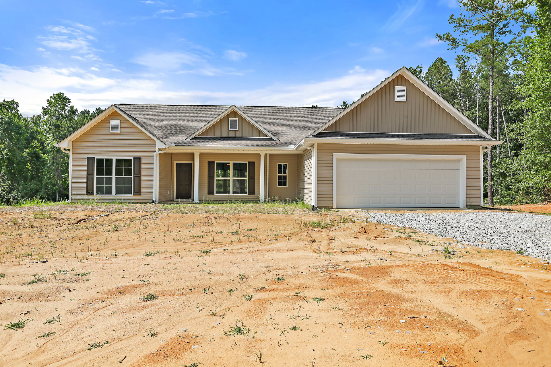 White garage door with brown trim, adjacent window with white frame, light-colored siding, dirt field in foreground, several white buildings in background, partly cloudy sky