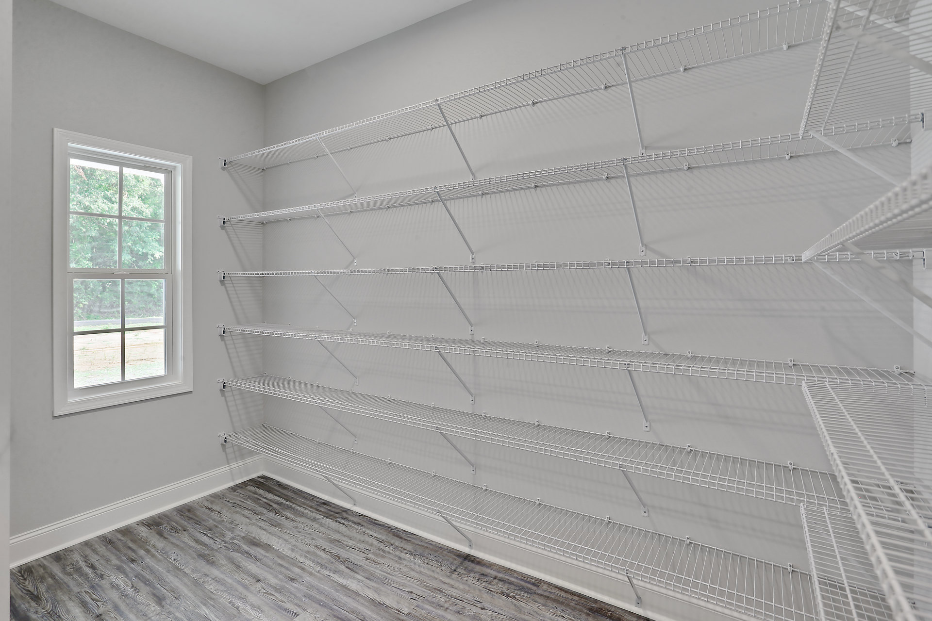 White built-in shelves with metal supports on a plaster wall, large window with daylight and view of trees, hickory flooring in a residential room.