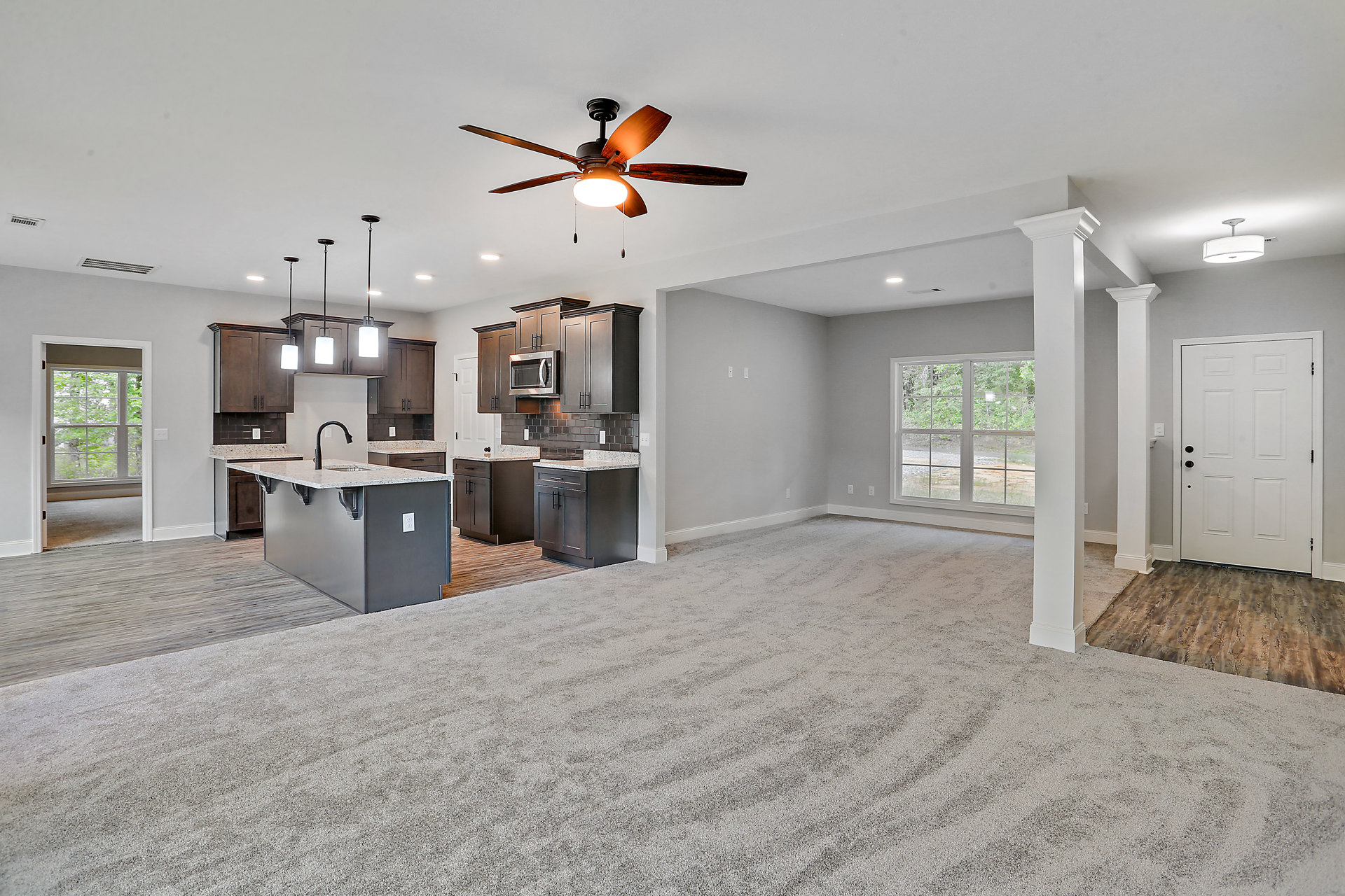 Open concept living room with wood flooring, ceiling fan with light, white door with black knobs, kitchen island featuring a sink, pendant light fixture, and window overlooking