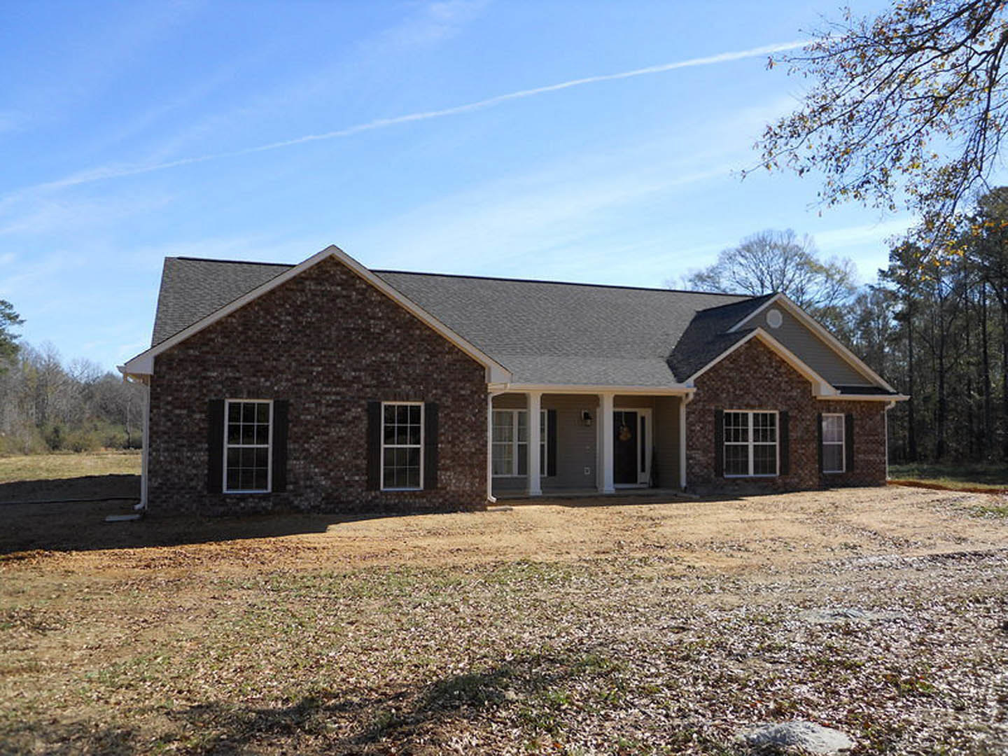 Red brick house with white roof, large paved driveway, white-framed windows, front door illuminated by exterior light, lawn scattered with leaves, trees and cloudy sky in