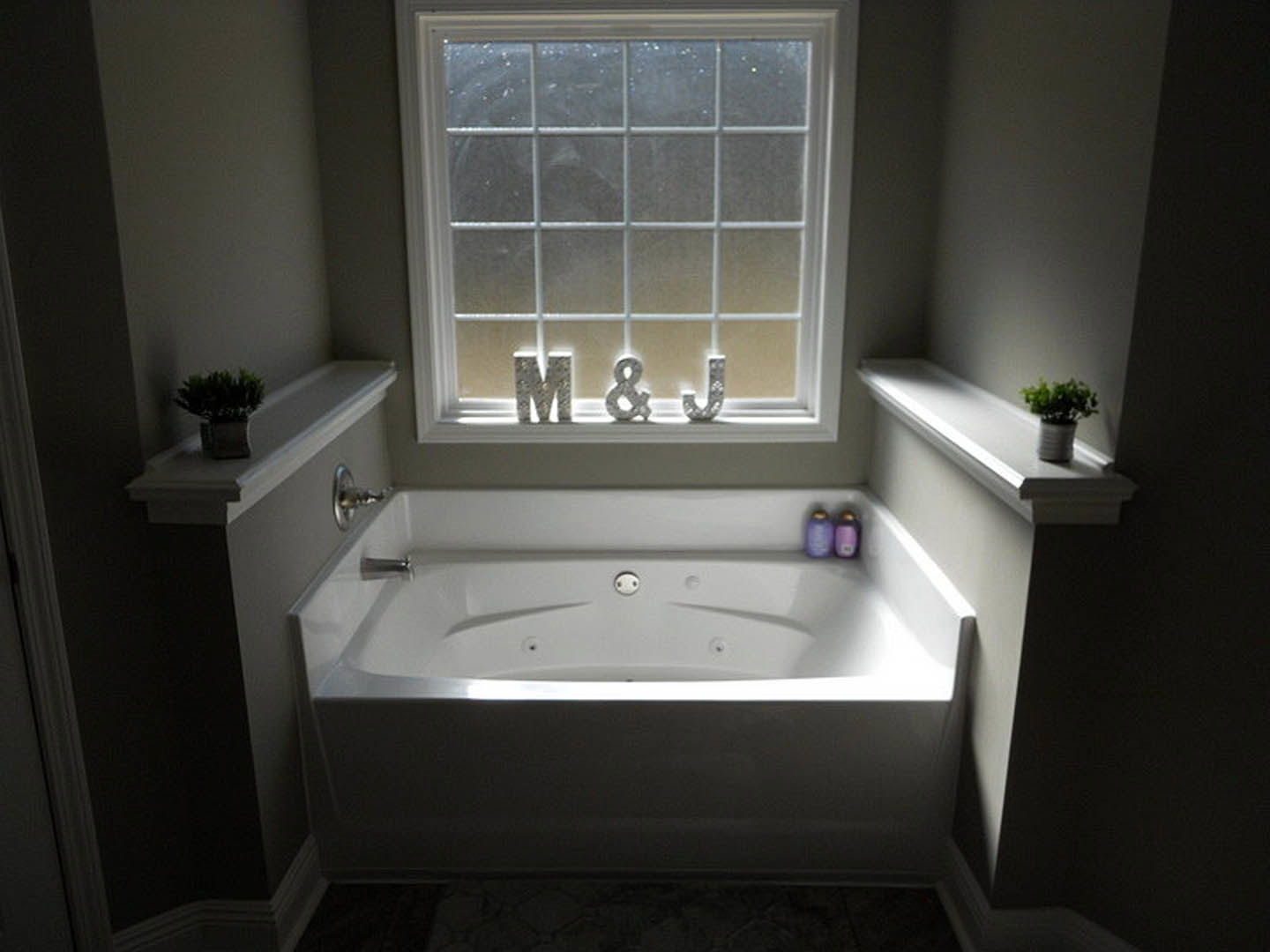 Freestanding white bathtub with silver trim beneath a large window, surrounded by light tile walls and a potted plant on the sill