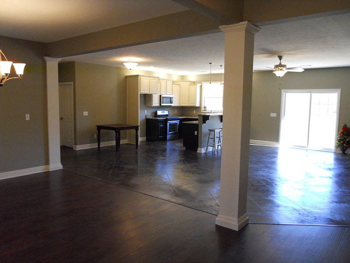 Open-concept living room with wood flooring, modern furniture, dining table, kitchen in background, large window letting in natural light, close-up of lamp and textured wall