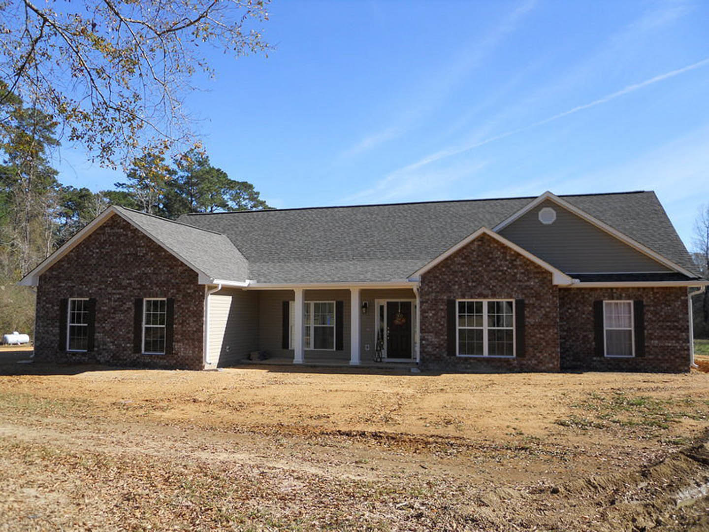 Red brick house with white-framed windows, covered front porch, dirt yard in foreground, tree branches and blue sky visible above