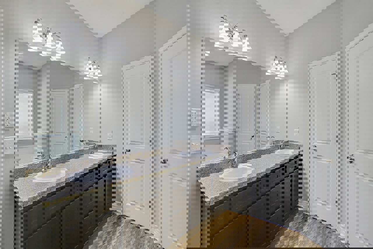 Bathroom with double sinks set in a light stone countertop, large frameless mirror above, white cabinetry below, silver faucets, wall-mounted light fixture with multiple lamps