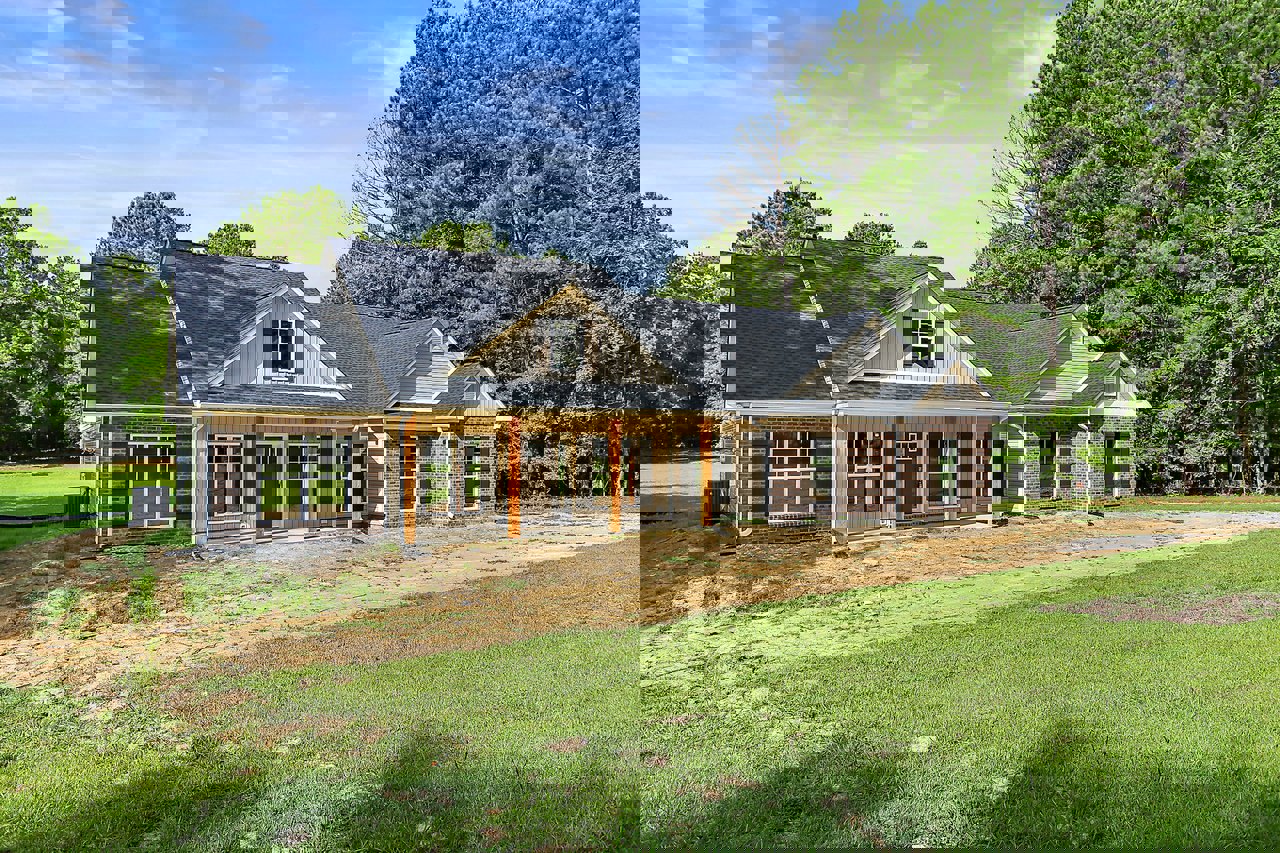 Two-story house with grey roof, white-framed windows, covered porch, green grass lawn, mature trees in background, partly cloudy sky