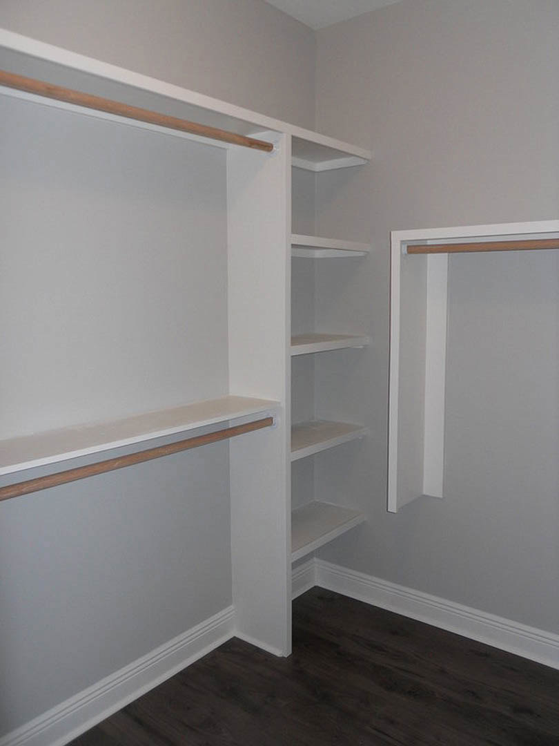 Closet with white shelves and wooden hanging bar, dark wood floor with white base trim, white walls, rectangular door centered in the space