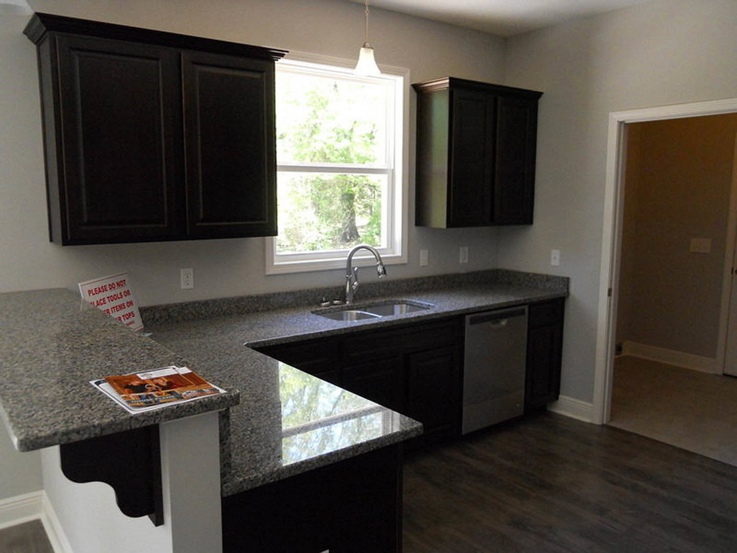 Granite countertops in a modern kitchen with black cabinets featuring gold edges, large window overlooking trees, magazine and sign on the floor