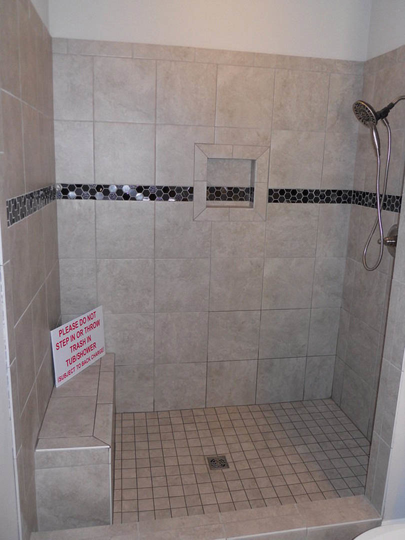 Walk-in tile shower featuring a built-in bench, handheld shower head with hose, marble tile flooring, recessed niche bordered in black and white tile, and a wall-mounted sign.