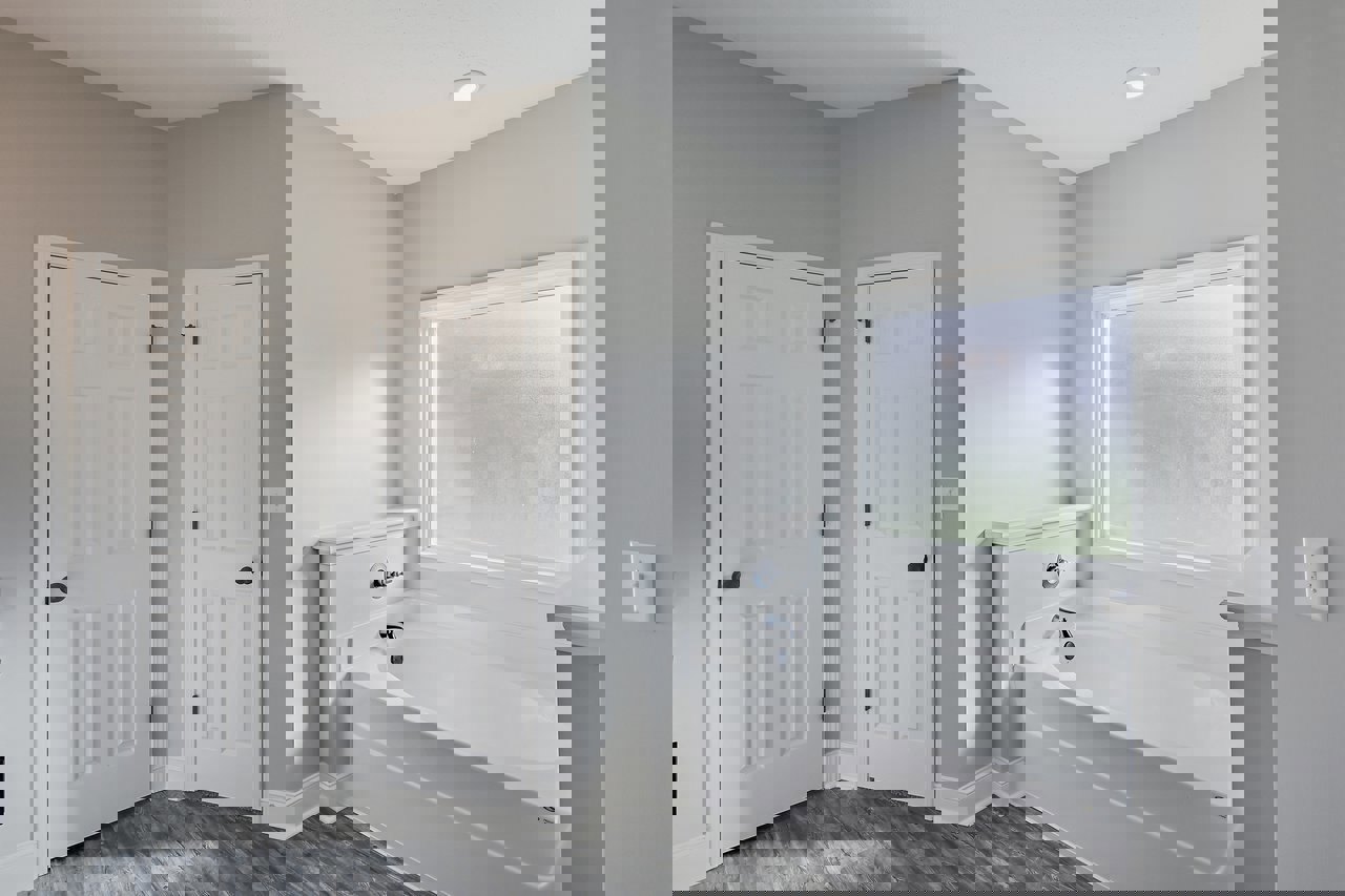 Bathroom with white freestanding bathtub, two white doors with black handles, frosted glass window, light switch on pale plaster wall, and light-colored flooring.