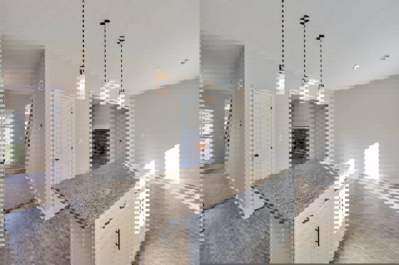 Marble kitchen island with white cabinets, stainless steel sink, white-framed window, brick fireplace wall, white door with black knob, light wood flooring, and black support pole