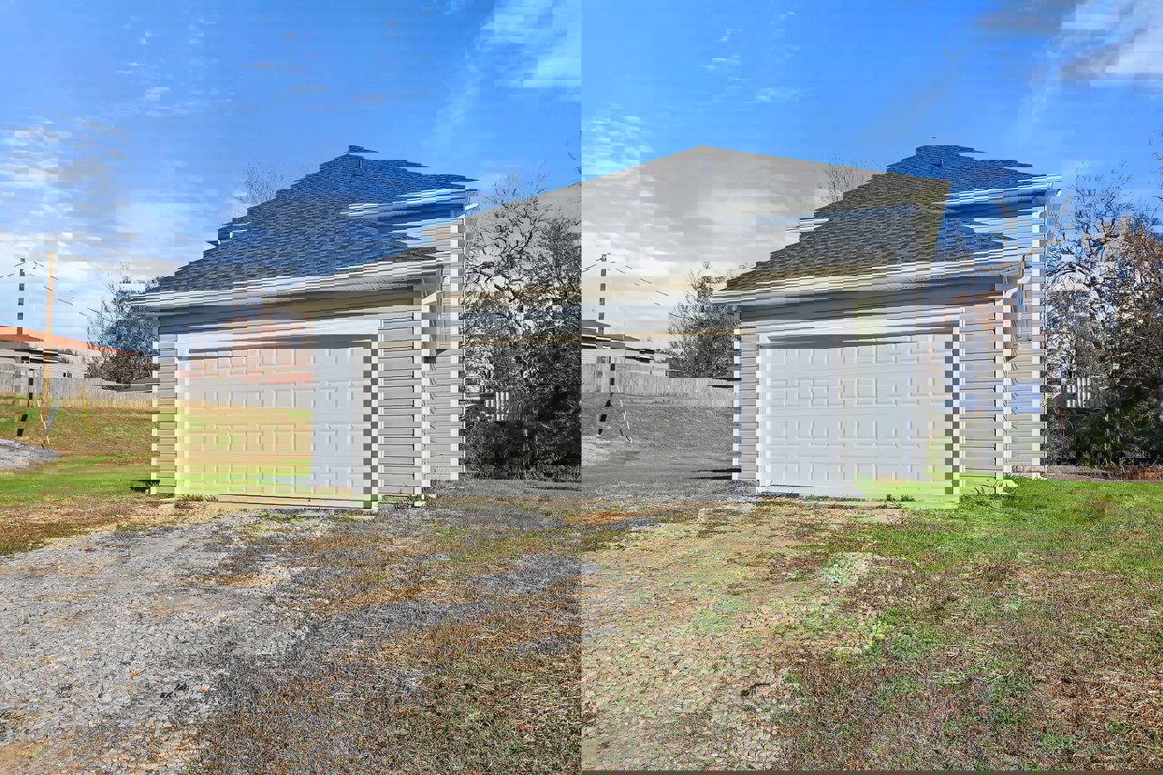 White garage door and roof on a house, gravel driveway bordered by grass and rocks, tree in yard under partly cloudy sky