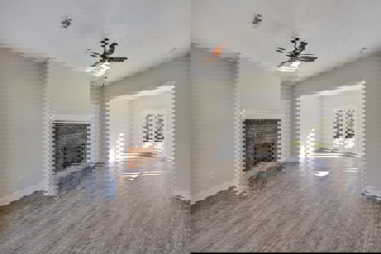 Living room with brick fireplace, wood flooring, ceiling fan with light, white double doors with glass panes, and ceiling vent