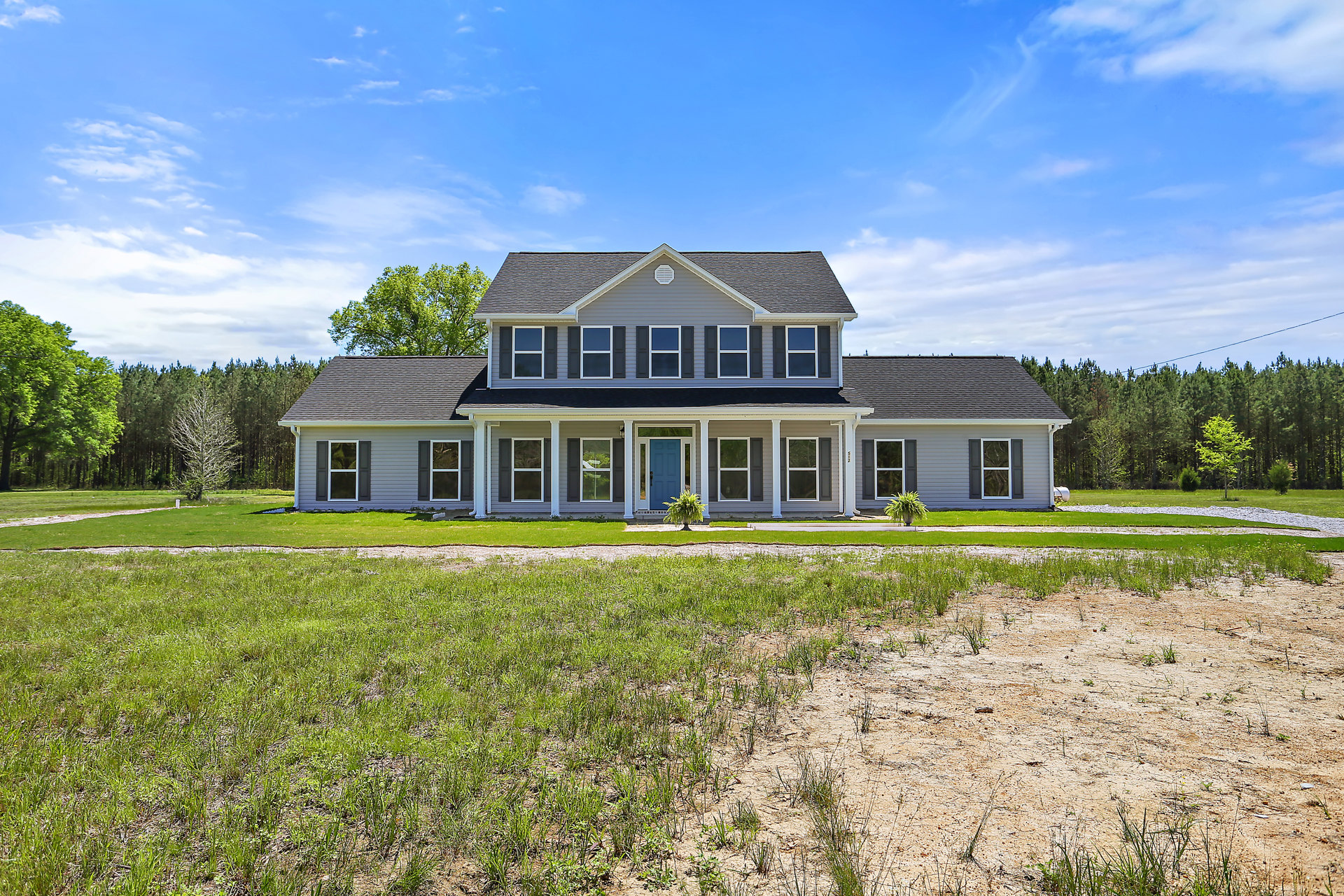 Two-story farmhouse with white siding, blue front door, white-framed windows, gray roof, expansive green lawn, and mature tree under a blue sky with scattered clouds