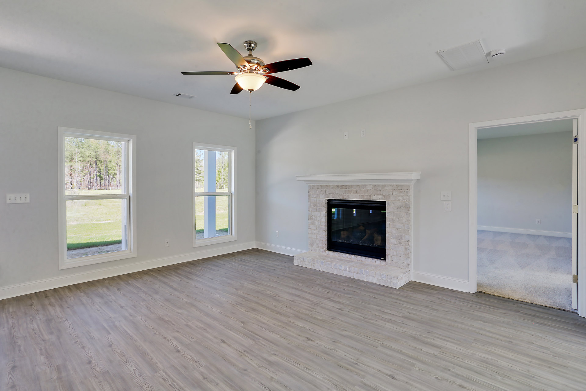 Living room with wood flooring, glass-door fireplace, ceiling fan with light fixture, large window showing trees outside