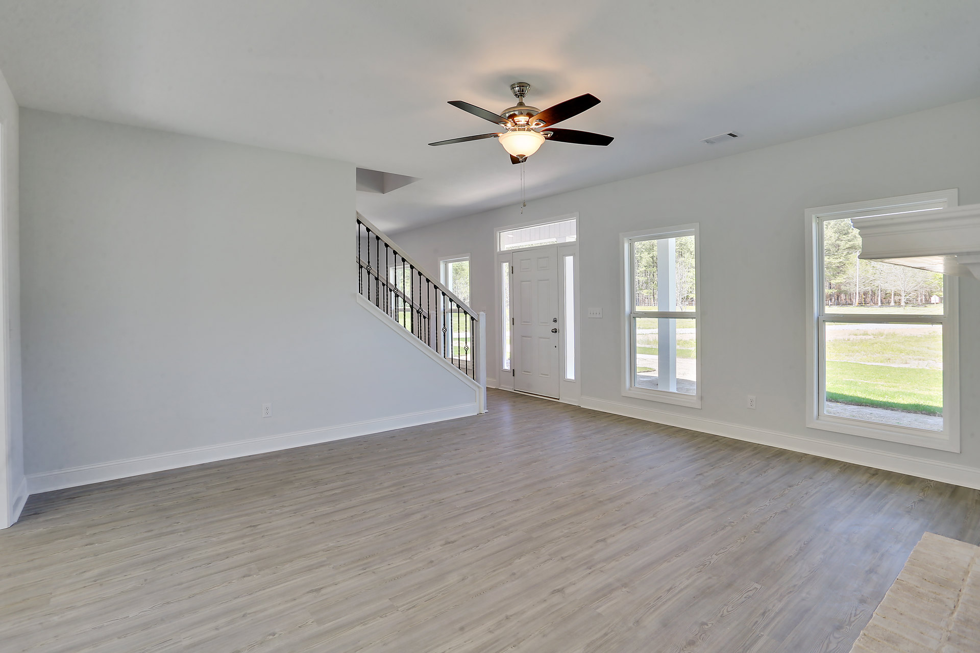 Hardwood floored room featuring a ceiling fan with light fixture, staircase with wooden railing, plaster walls, and large window overlooking trees.