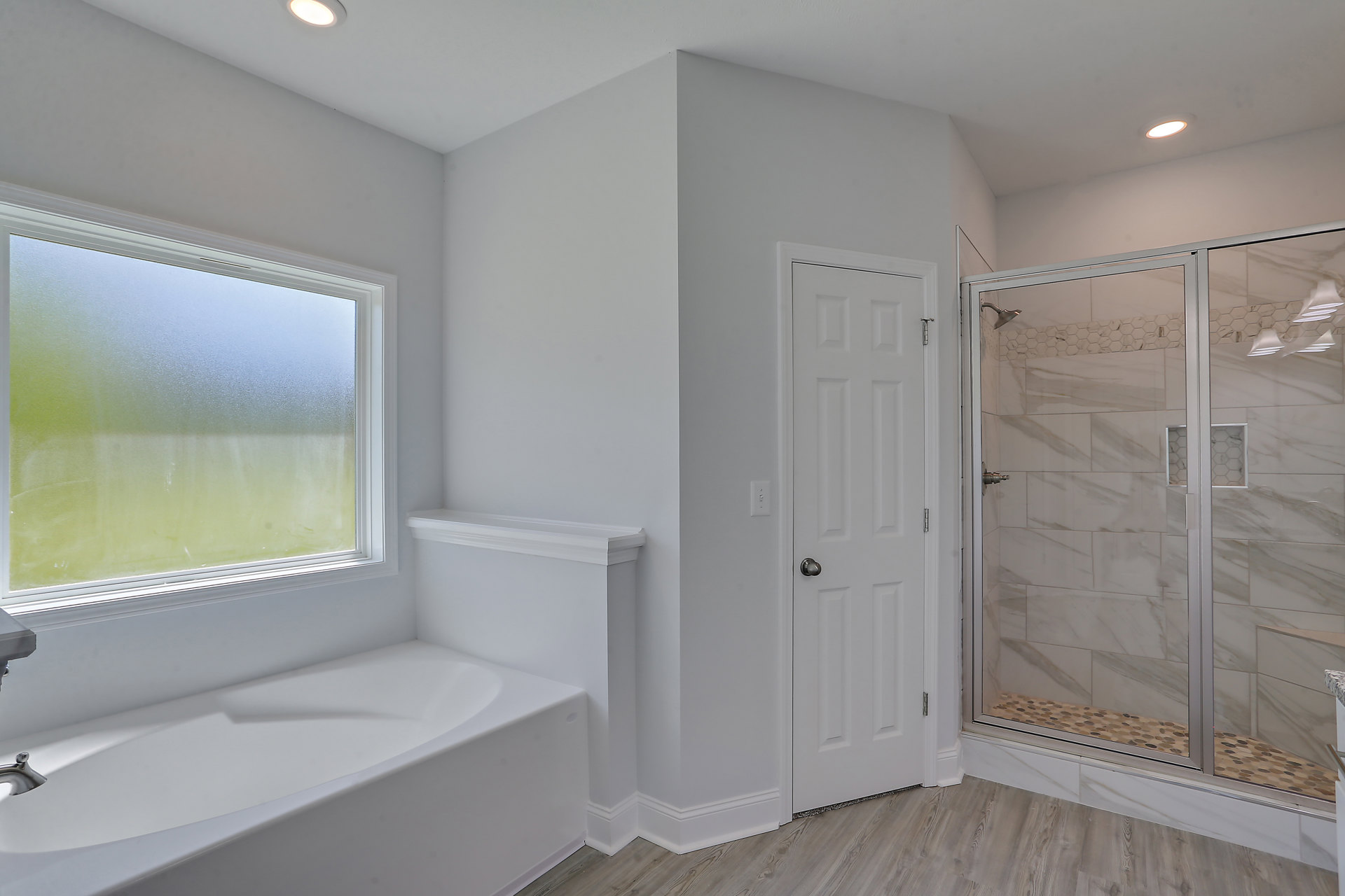 Bathroom with white tile floors, glass shower enclosure, freestanding bathtub beneath frosted window, silver fixtures, and white door with chrome handle