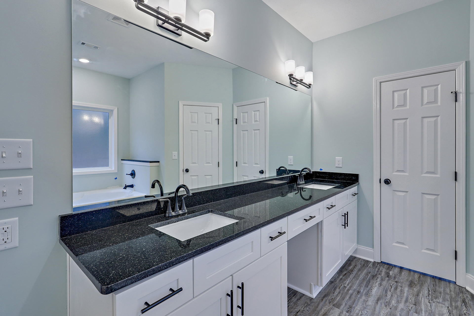 Double vanity bathroom with black countertop, two white sinks, large wall mirror, white cabinetry, and white door with silver doorknob.