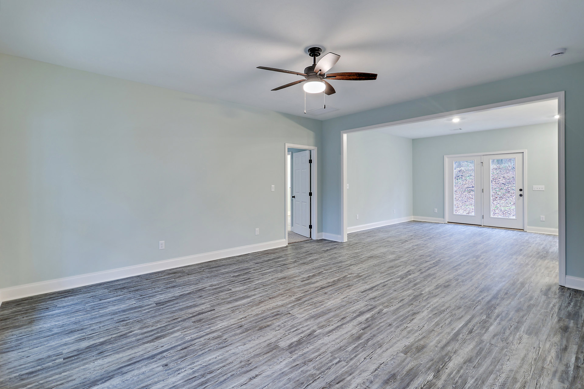 Wood floor room with white walls, ceiling fan with light, double glass-paneled doors, and sunlight streaming across the floor.