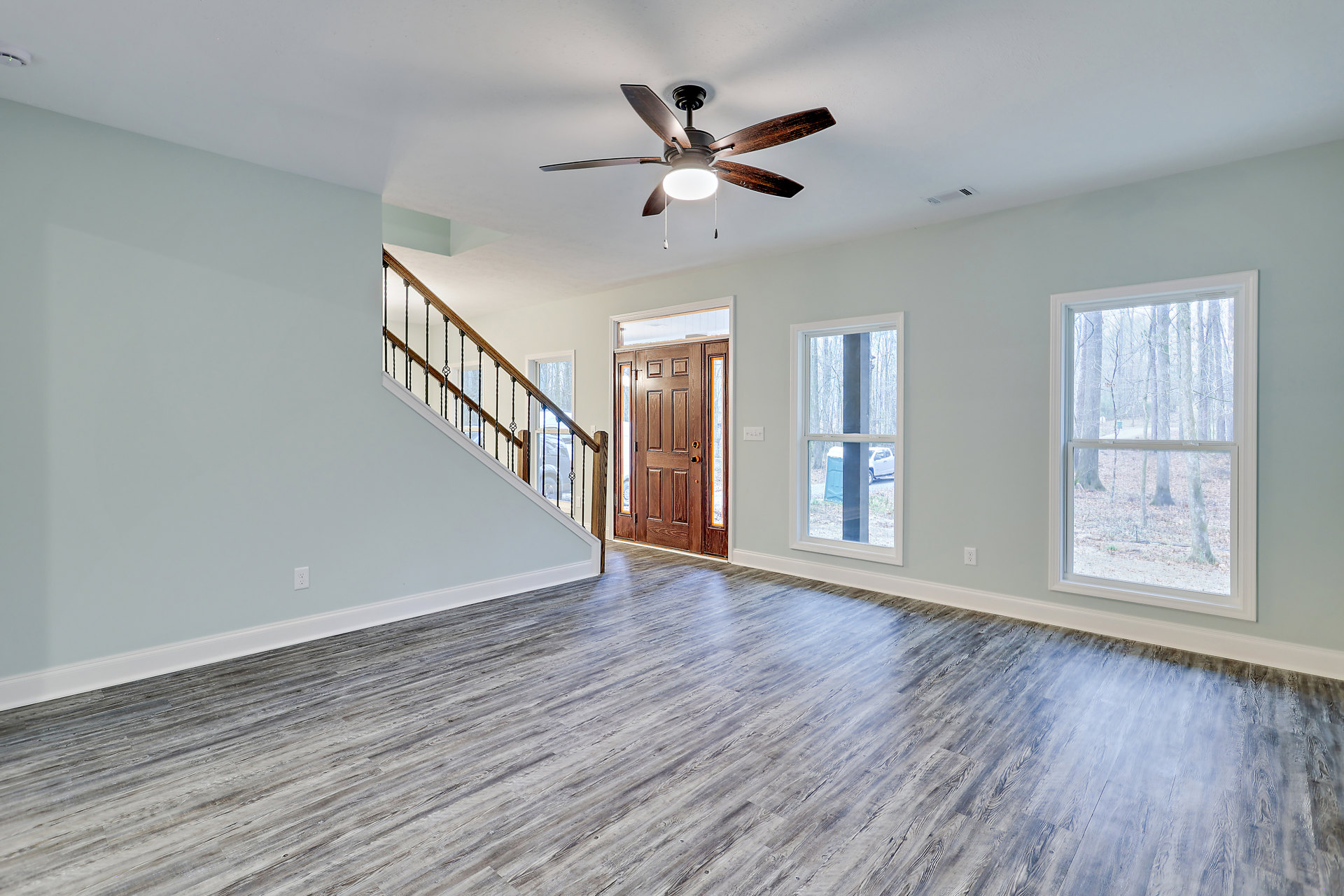 Open living area with wood flooring, ceiling fan with light, metal-railed staircase, brown door with glass panel, large window showing trees outside