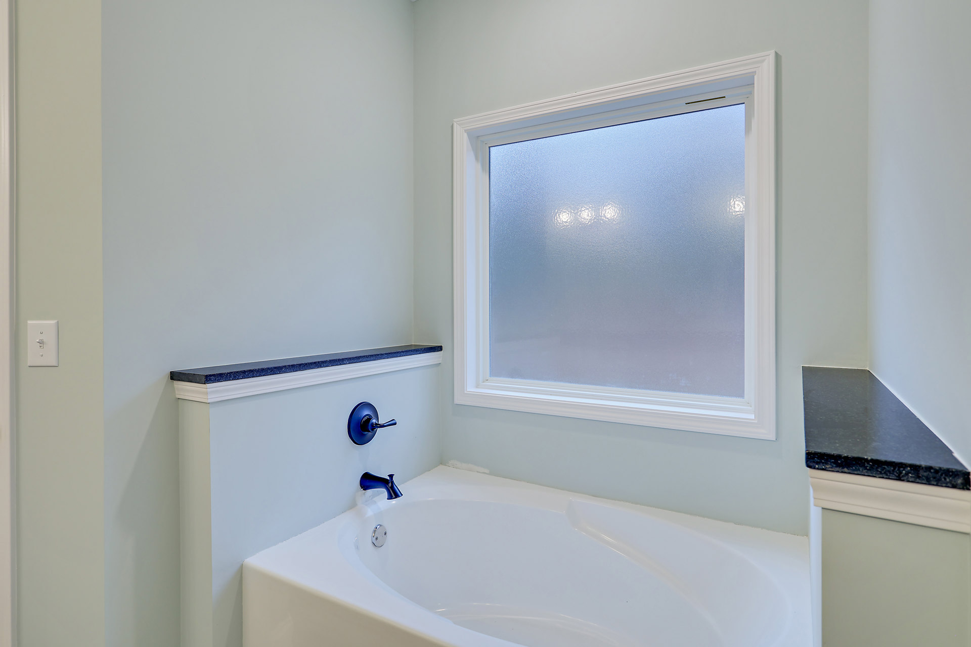 Freestanding white bathtub beneath frosted window, white tile walls, chrome faucet, light switch on adjacent wall