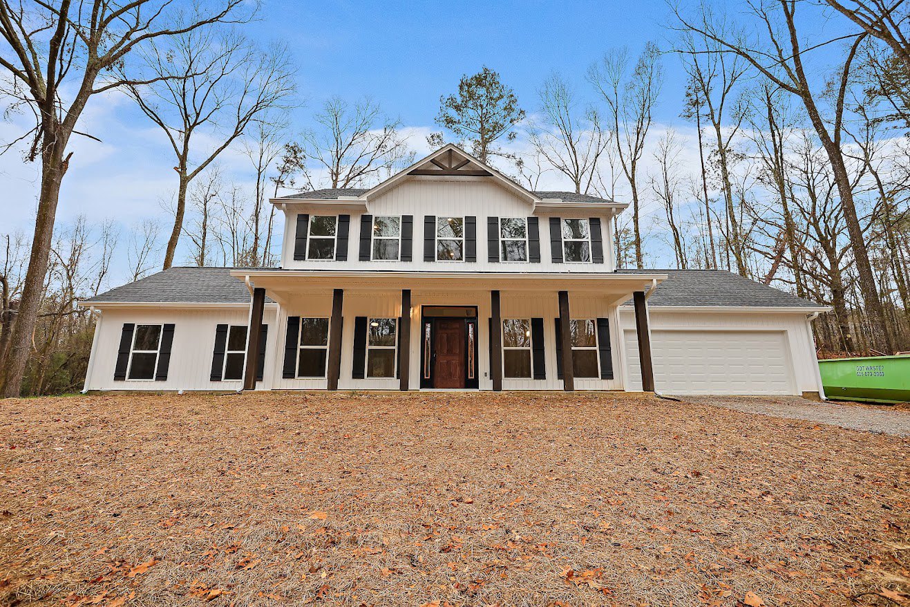 White two-story house with brown wooden front door, wide driveway, manicured lawn, leafy trees in background, and green waste container near porch