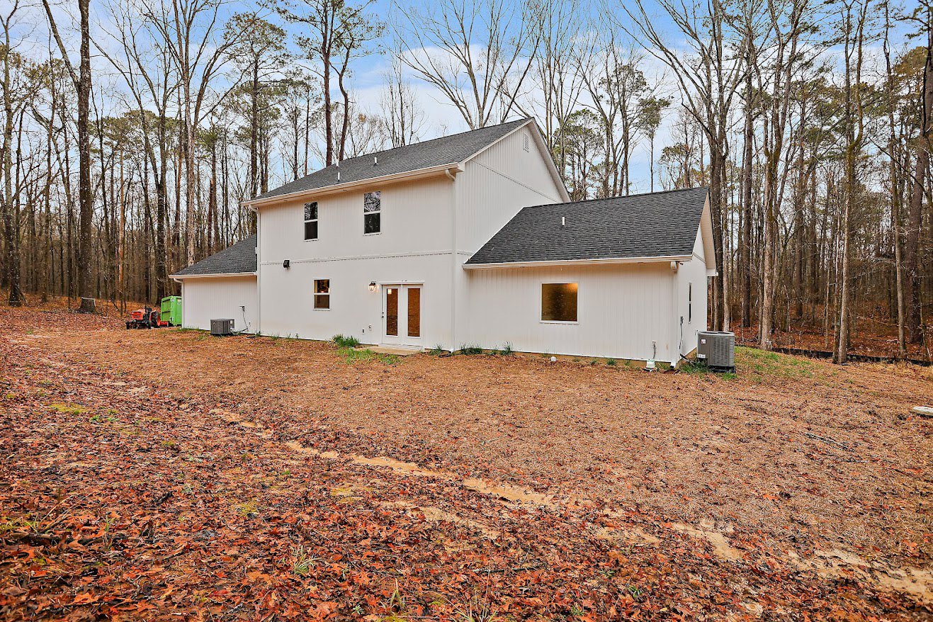 White siding house with gabled roof, large windows, brown leaves scattered on ground, surrounded by mature trees under clear sky