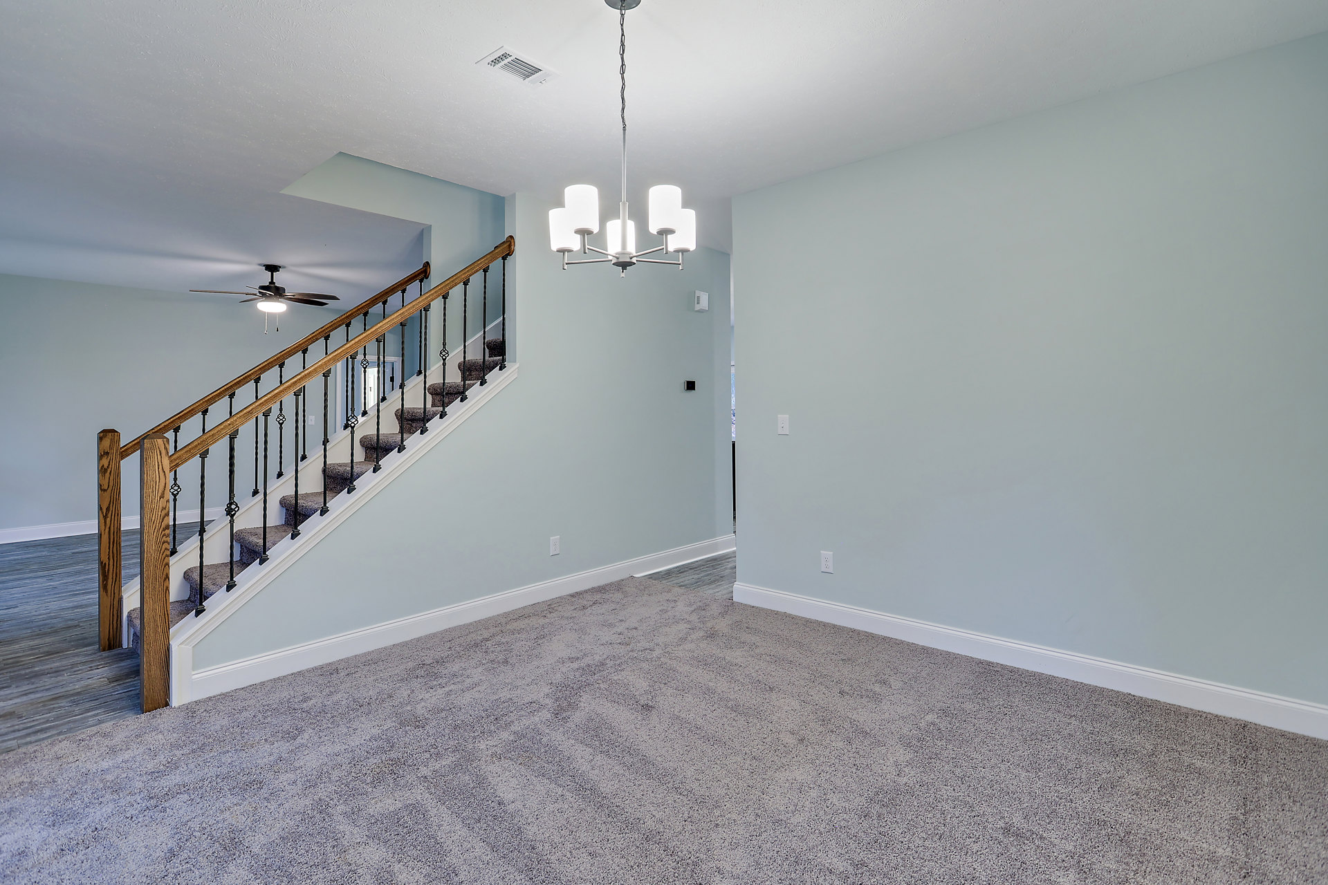 Open living area with plaster walls, wood flooring, central staircase featuring metal handrail, ceiling fan with light fixture, and recessed ceiling vent.