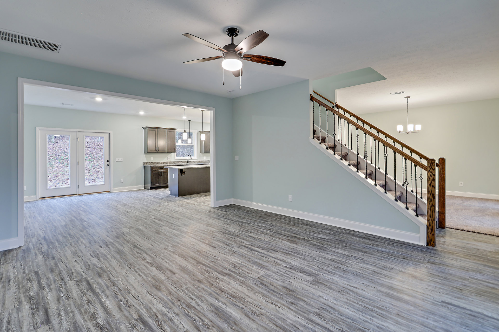 Open living area with wood flooring, white walls, glass-paneled double doors, staircase with dark wood railing, ceiling fan with light fixture, and built-in cabinetry