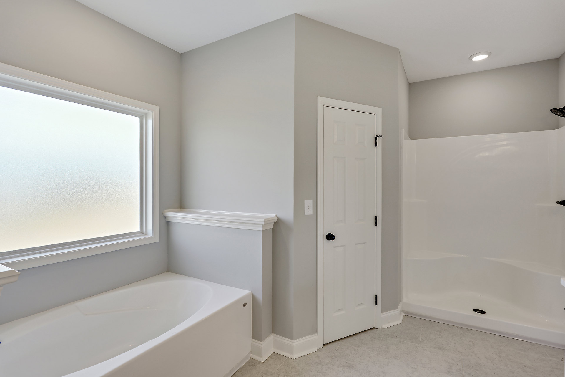 White bathtub next to glass-enclosed shower with black handle, frosted window above, white walls and tile, white door with black knob