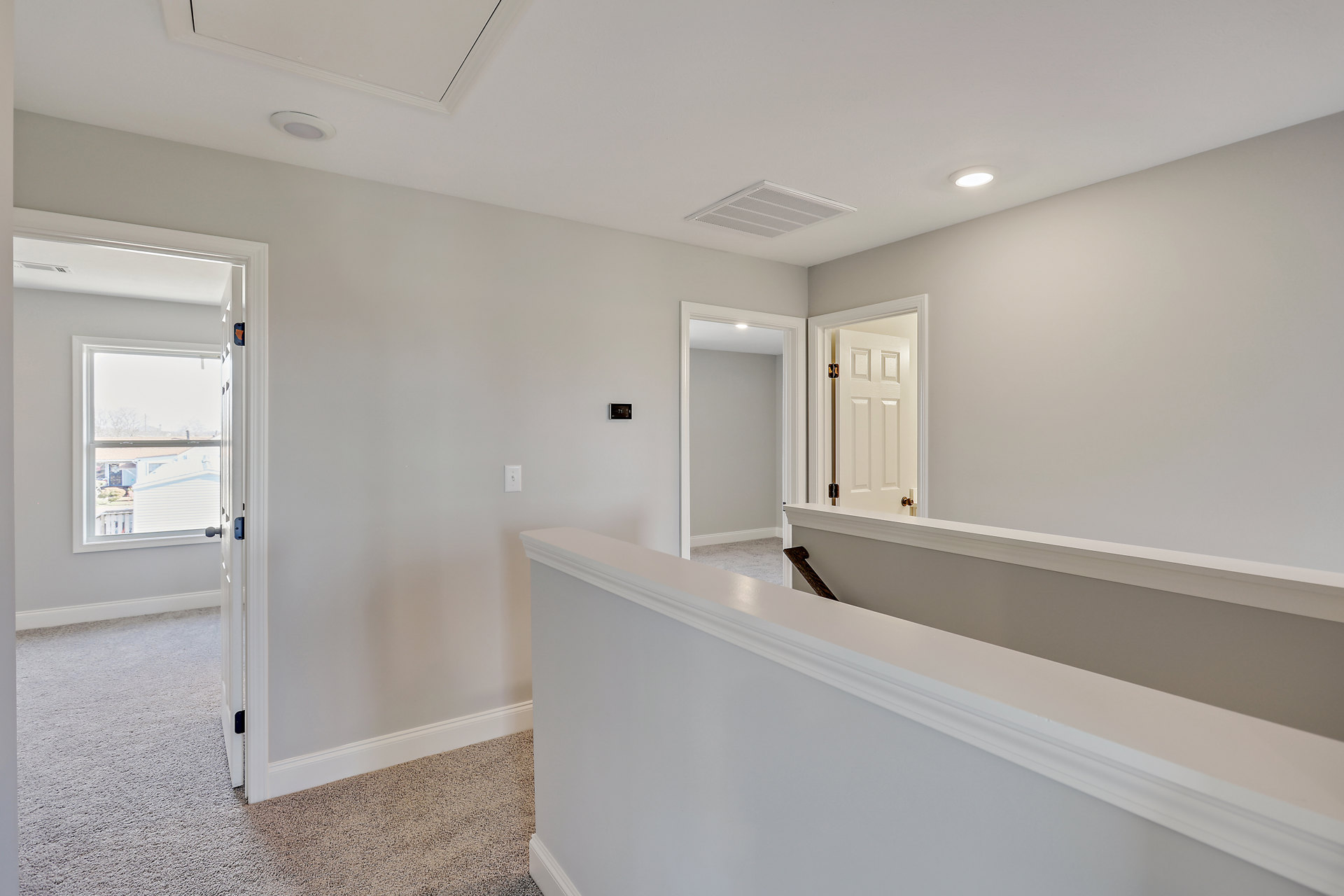 Hallway with white painted walls, white door with visible hinge, white ceiling vent, white railing, tile flooring, and window overlooking neighborhood houses
