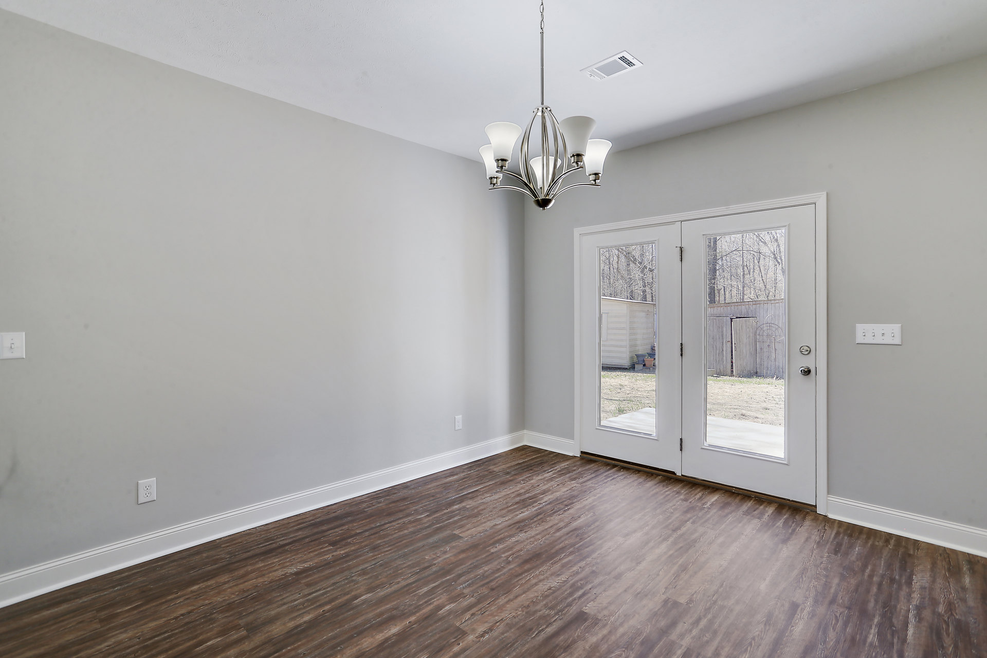 Wood flooring in a spacious room with glass-paneled double doors, white walls, and a decorative chandelier hanging from the ceiling