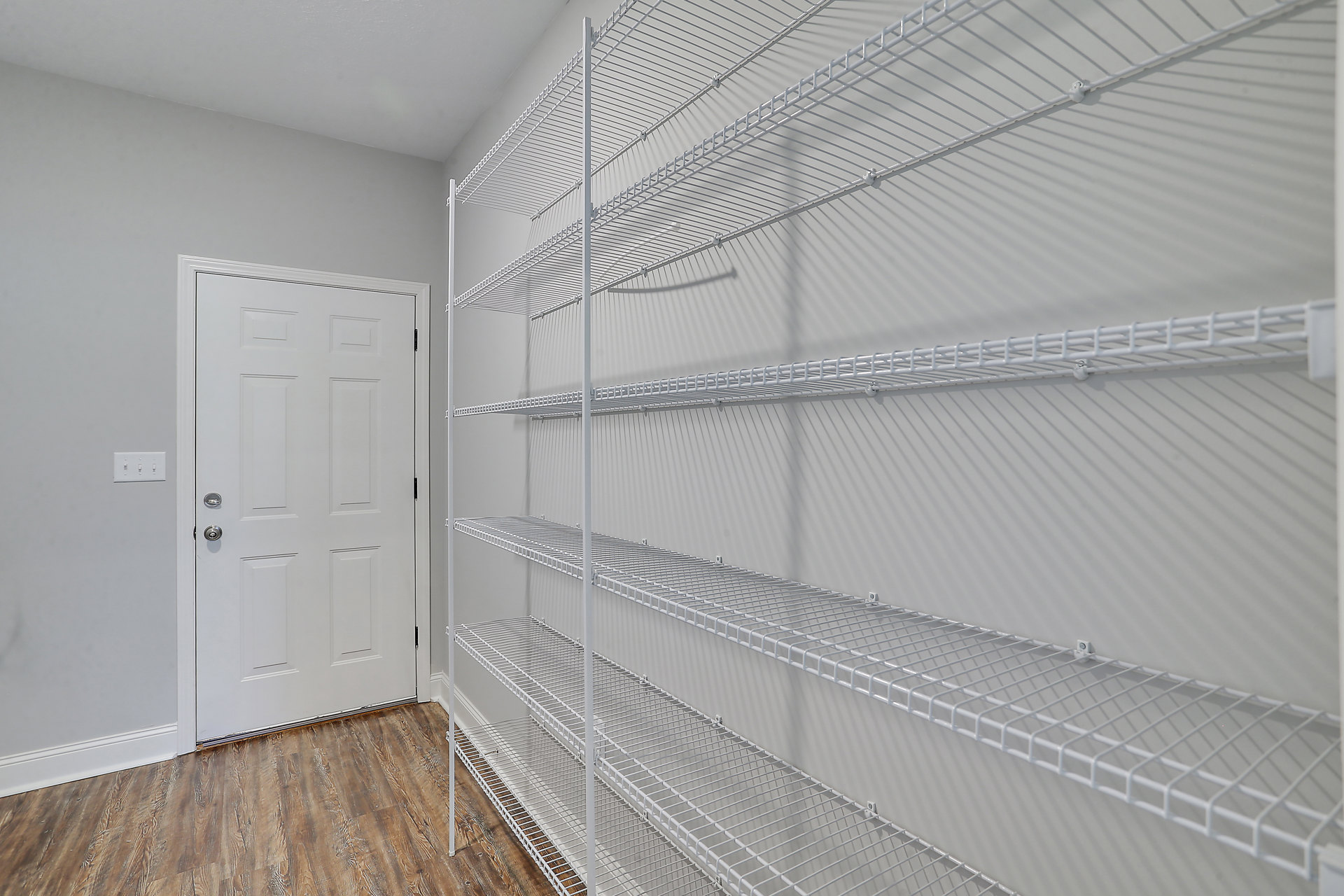White wire shelves mounted on a plaster wall, white door with silver handle, wood flooring, and smooth white ceiling in a residential interior.