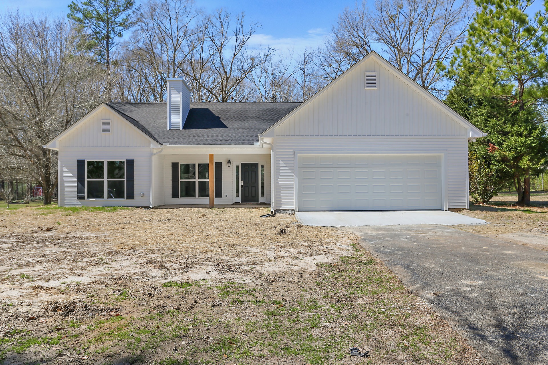 Two-story house with white siding, black front door, white-framed windows, attached garage, paved driveway, mature trees in the background, and a small white cottage visible
