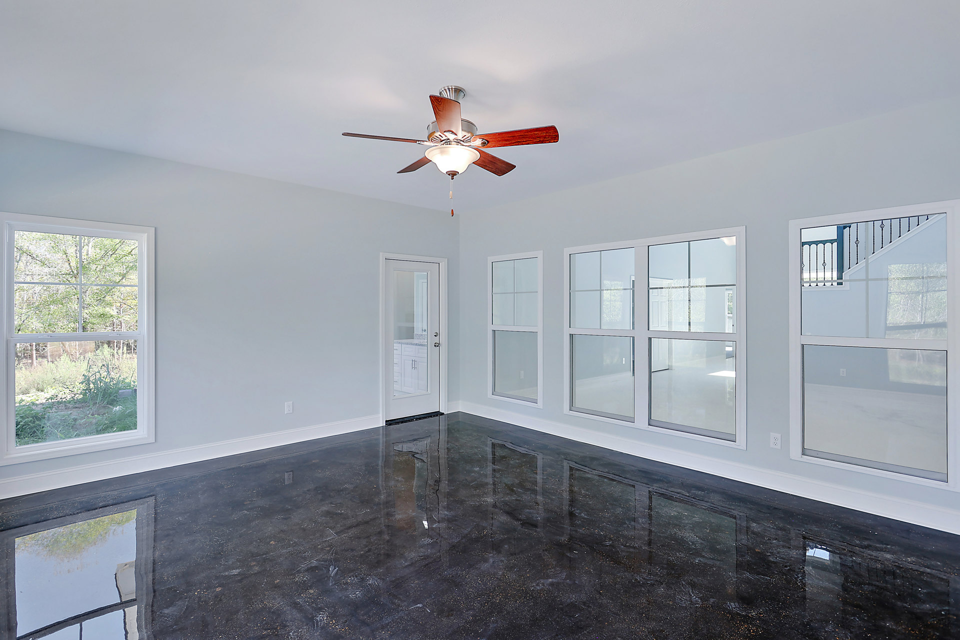 Ceiling fan with light fixture mounted on white ceiling, shiny black floor, white walls, large window showing trees outside, glass door with metal railing, white door adjacent to