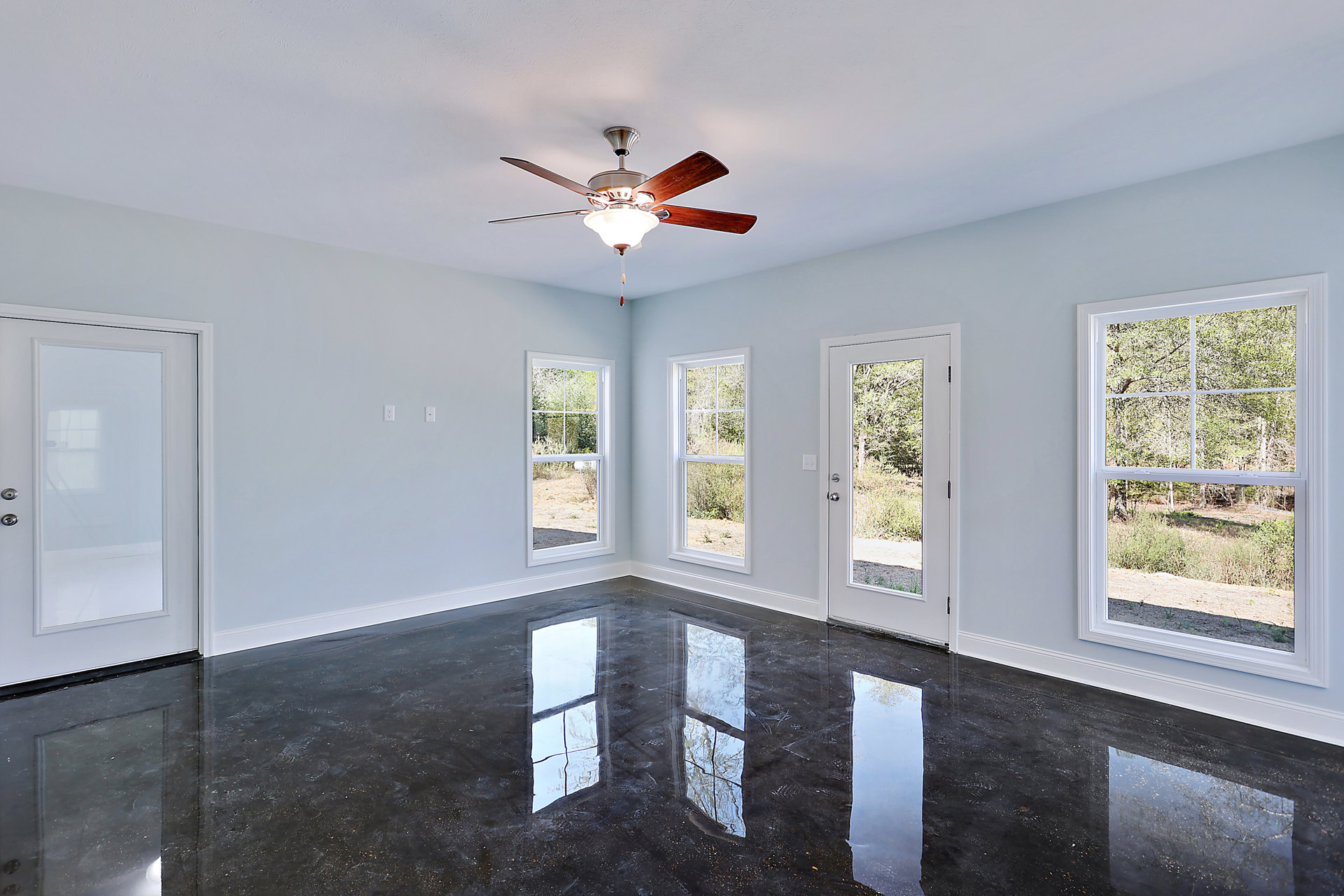 Ceiling fan with light fixture mounted on white ceiling, shiny hardwood floor reflecting sunlight from multiple large windows, white walls, glass-paneled door, and view of green