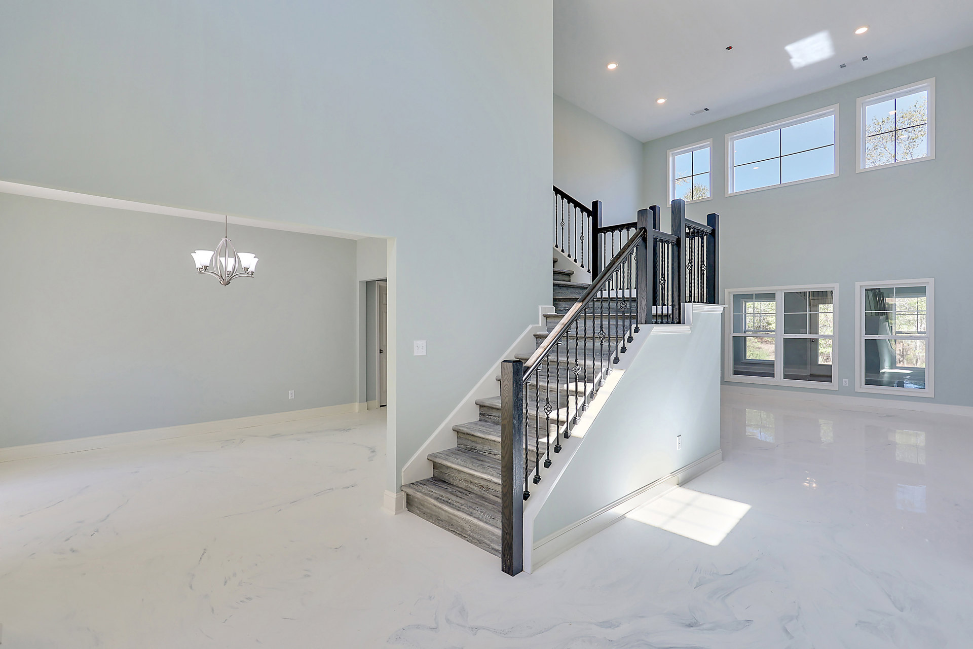 Wood staircase with black metal railings, white plaster walls, large window framed in white revealing leafy tree outside, modern chandelier hanging from ceiling, light hardwood