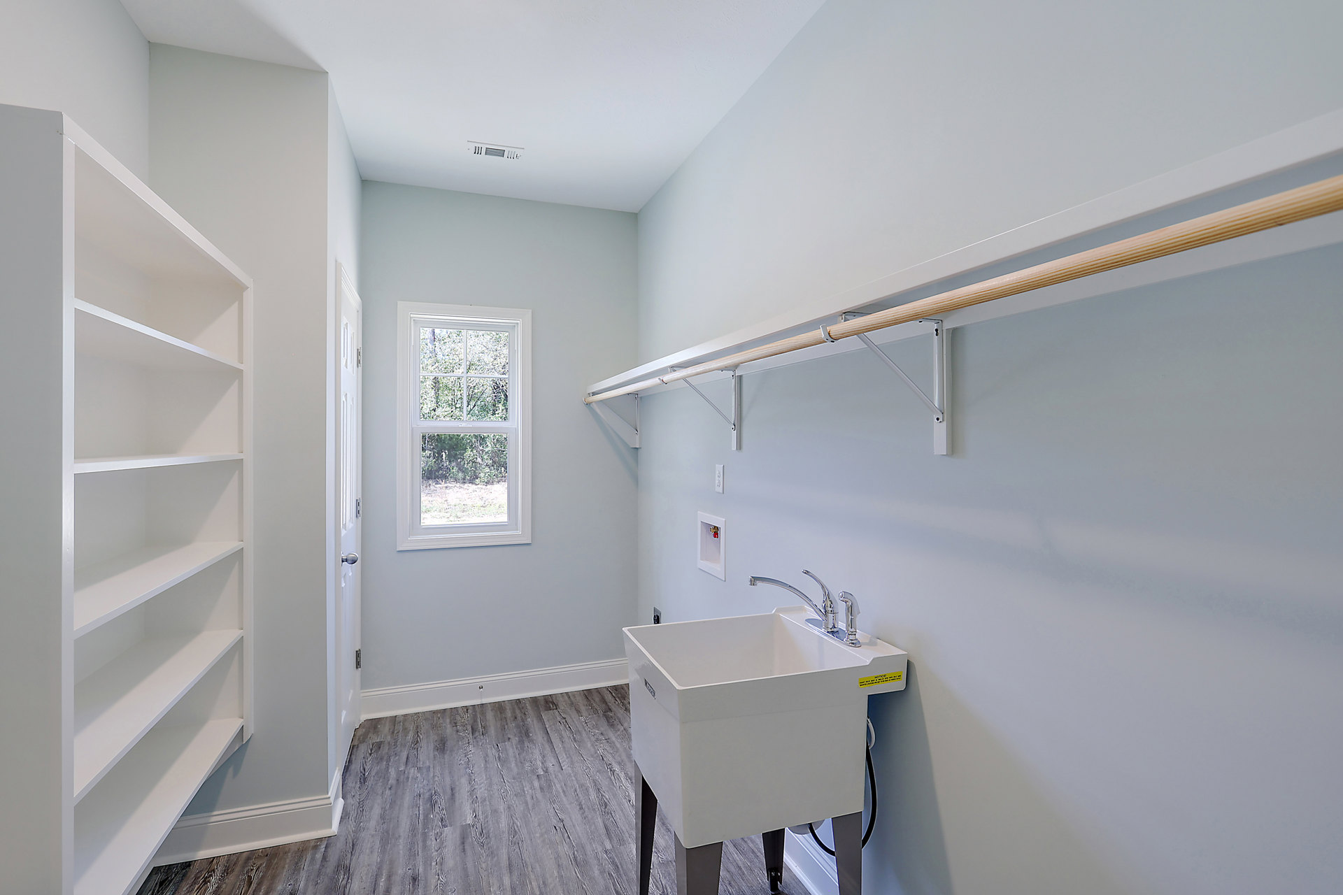 Laundry room with white cabinetry, open shelves, built-in sink with silver faucet, window overlooking trees, light-colored walls and flooring